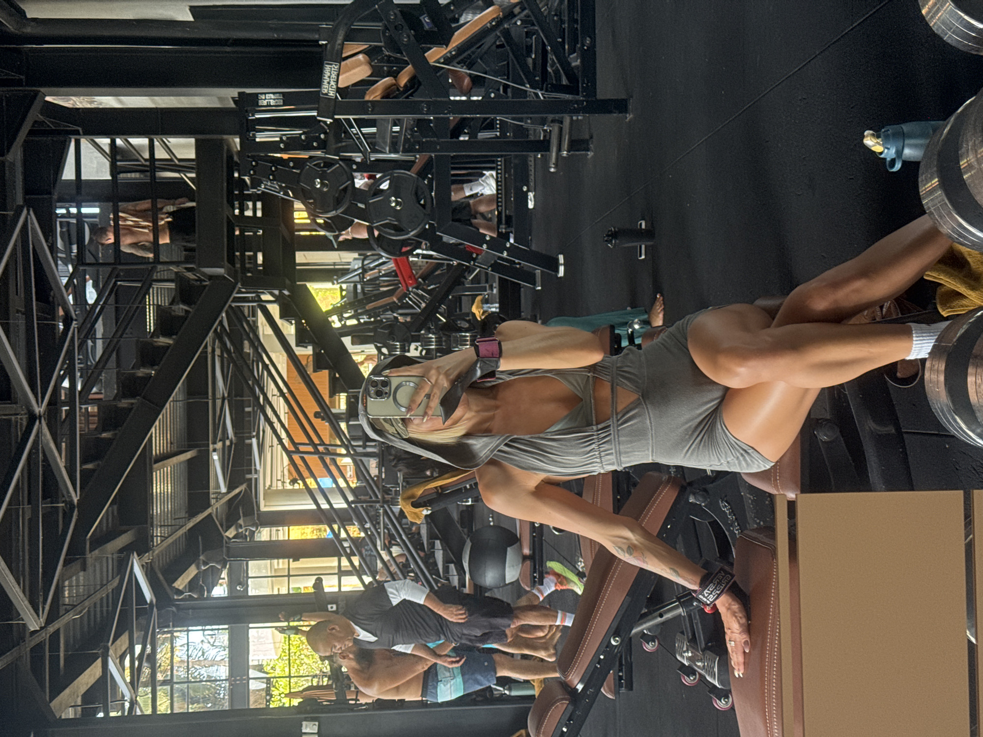 In a brightly lit, modern gym in Ubud District, Indonesia, a woman with blonde hair is taking a selfie in the mirror. She is wearing a grey hooded athleisure set consisting of a halter top and shorts. She is sitting on a brown leather bench, with her right arm resting on the bench and her left hand holding her phone. She is wearing a watch on her left wrist and wrist wraps on both wrists. In the background, other gym-goers are visible, some exercising and others walking around. The gym features industrial-style architecture with metal stairs and beams, along with various weightlifting machines and equipment. Sunlight streams in from large windows, illuminating the space.