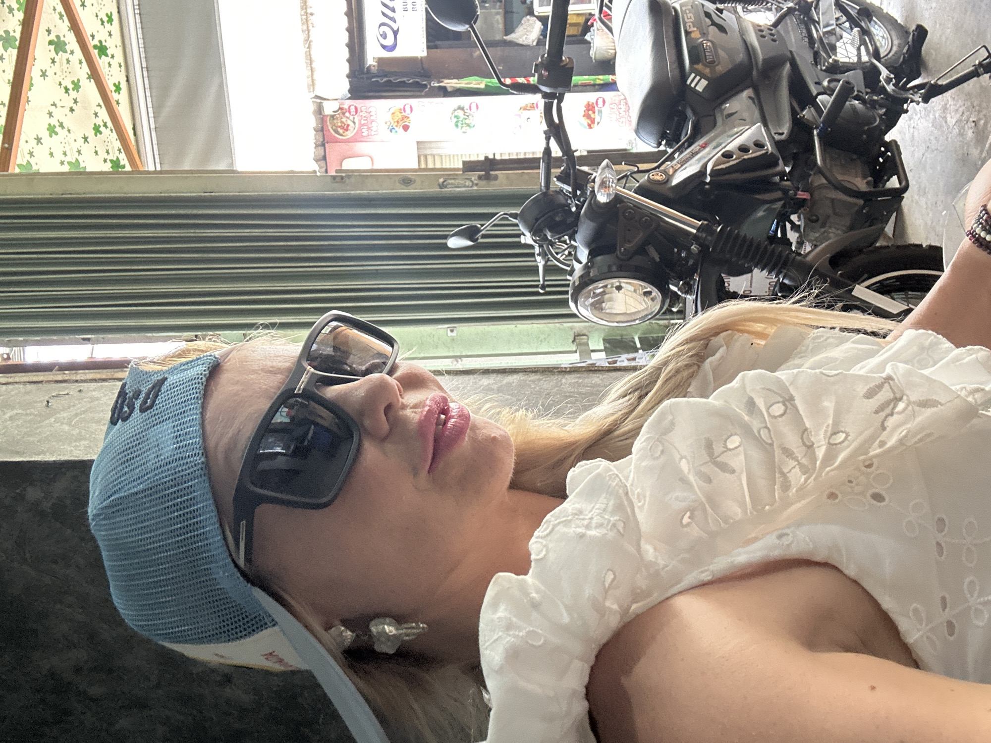 A woman wearing a blue mesh cap, sunglasses, and a white blouse is positioned in front of a closed, horizontally corrugated metal security gate, likely a storefront shutter. A black motorcycle with a top case is located behind her and to the side. The background reveals storefronts with signage written in Vietnamese, and the style of the signage indicates that the location is likely Nha Trang, Vietnam.