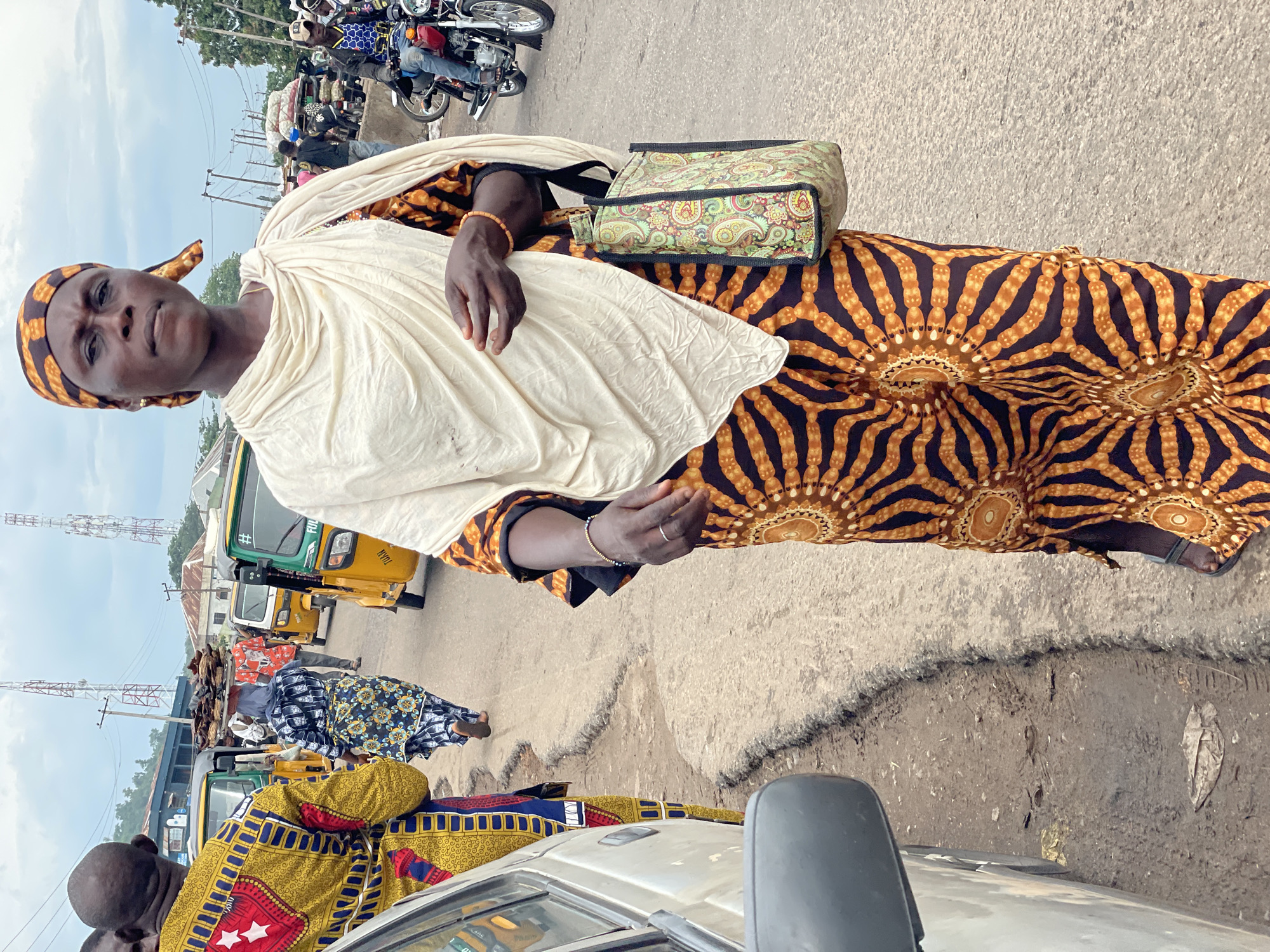 The image captures a street scene in Lokoja, Nigeria, likely during the day with overcast skies. The primary subject is a woman standing in the foreground, wearing a vibrant orange and brown patterned dress and a cream-colored headscarf. She has a patterned bag slung over her shoulder. Her expression appears serious or contemplative.
The background is filled with activity, suggesting a busy marketplace or transportation hub. Several auto-rickshaws (Keke NAPEPs), a common mode of transport in Nigeria, are visible, some appearing to be yellow and green. A few motorcycles are also present, with riders. Other people are seen in the background, some walking and others seated or standing near the vehicles. The ground is a mix of asphalt and dirt, with some unevenness and debris. Power lines and utility poles are visible in the distance, and there are some buildings and vegetation.
The overall impression is one of daily life and commerce in an African urban setting. The bright colors of the clothing and vehicles contrast with the somewhat muted tones of the road and sky. The scene is likely a roadside area where people are waiting for transportation or conducting business. There is no discernable text in the image.