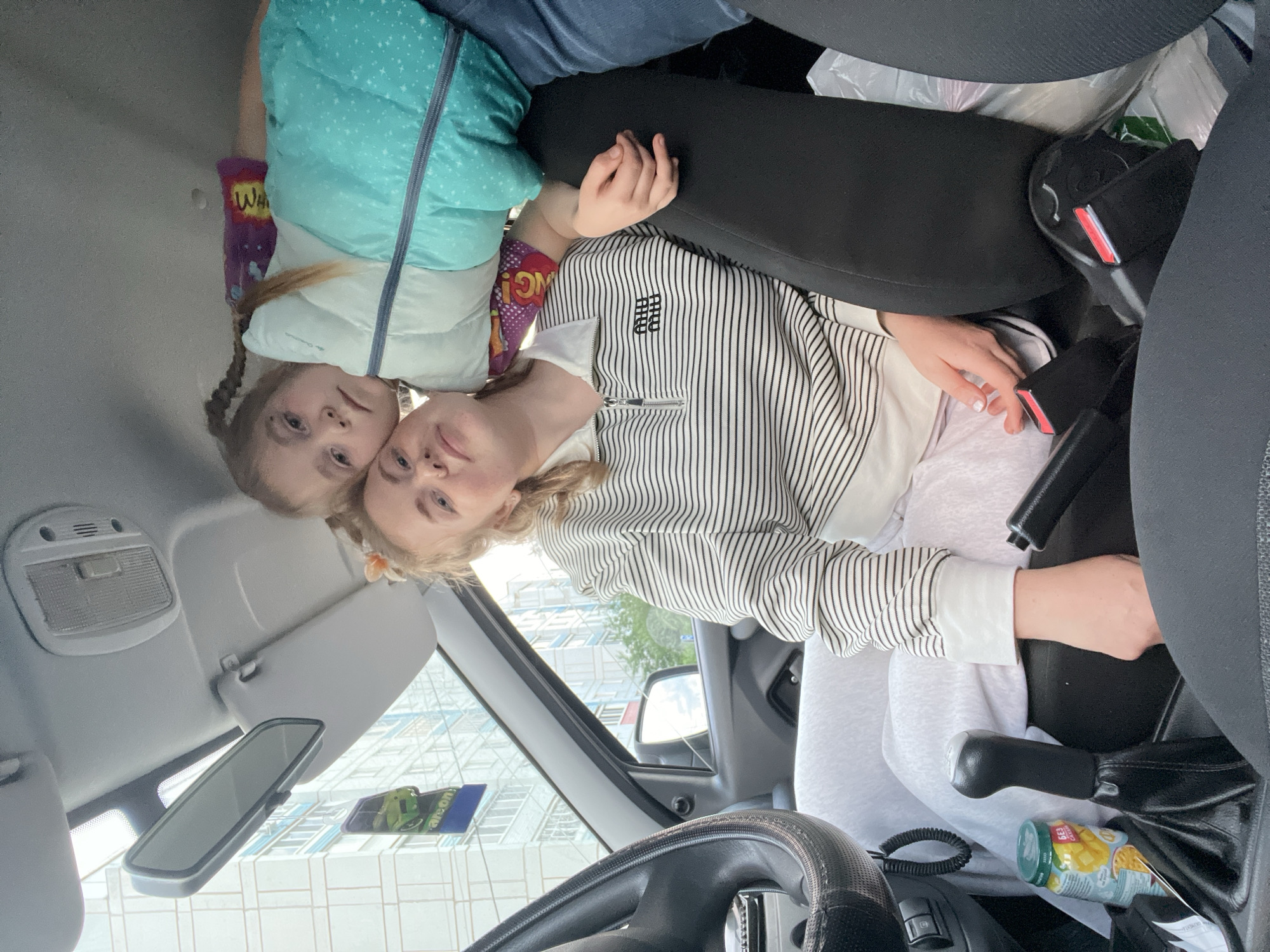 Two females are inside a car. One female with blonde hair and wearing a striped shirt is in the driver's seat. The other female, a young girl, is beside her wearing a turquoise jacket and has blonde braided hair. The car's interior is visible, including the steering wheel, gear shift, and rearview mirror. Outside the car window, a multi-story building is visible. The image was taken in the city of Chaikovsky, Russia.