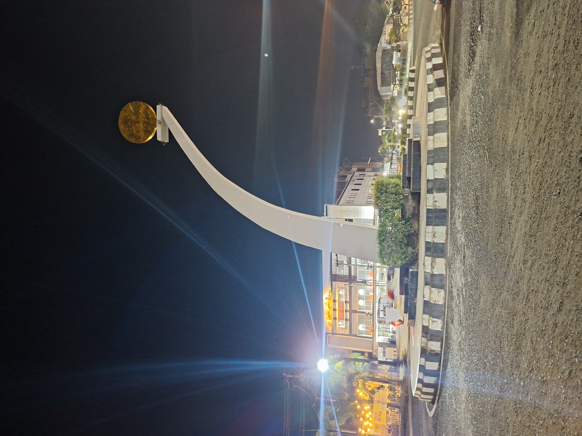 The image shows the Tugu Simpang Jam monument in Lhokseumawe, Indonesia, at night. The monument features a curved white arm topped with a golden disc. The background shows buildings, streetlights, and some vegetation. The foreground consists of a section of road with striped curbs. The scene is illuminated by streetlights, causing lens flare. No people are clearly visible.