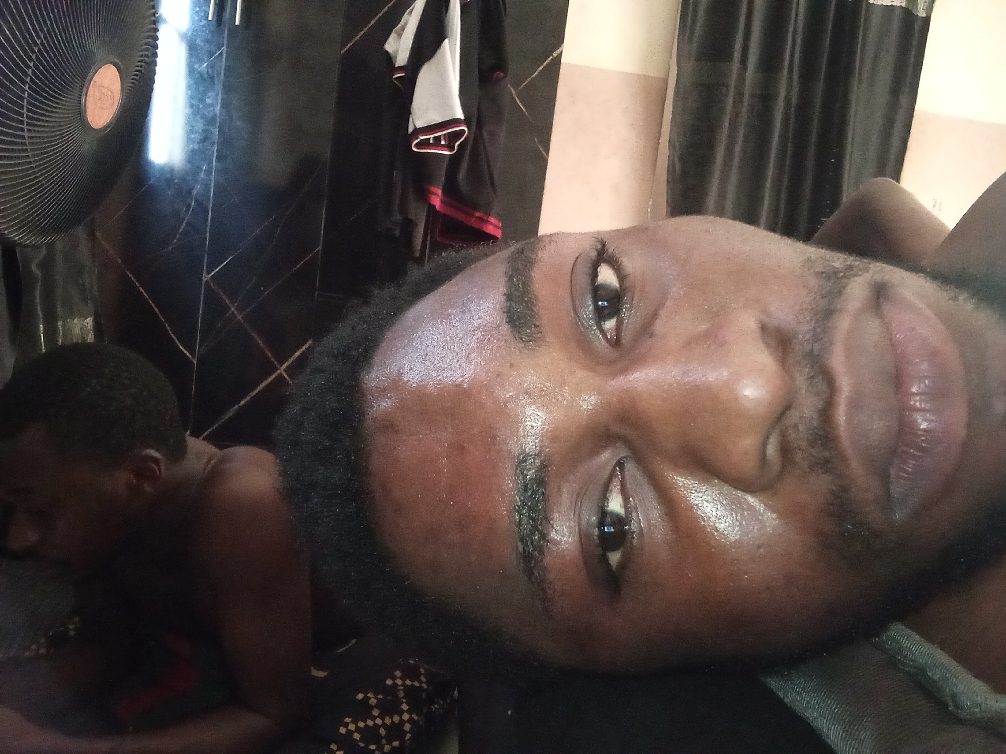 The image shows a close-up of a person lying down, with another person in the background to the left. The primary subject's face is tilted upwards, with their eye visible. They have dark skin and appear to be sweating. Their lips are slightly parted, and they have a light beard. In the background, a fan is visible on the left, and dark curtains are on the right. There are also clothes hanging on a rack in the background. The scene is indoors. The location is Lokoja, Nigeria.