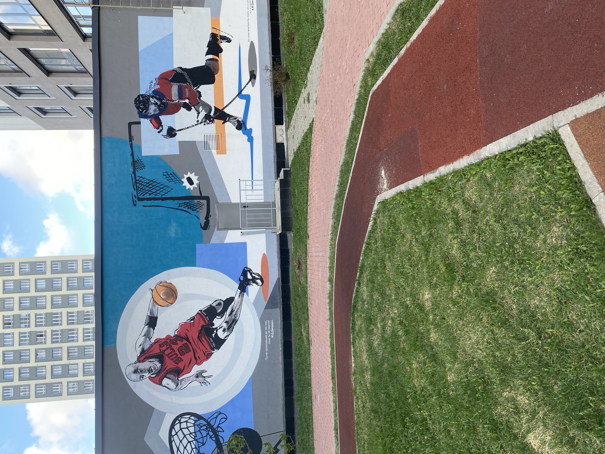 The scene is a vibrant mural on the side of a building in Yekaterinburg, Russia. The mural depicts two sports: basketball and hockey. On the bottom half, a basketball player in a Bulls jersey is shown dunking, while the upper half shows a hockey player with a stick. Nearby is a red and brick-colored pathway, surrounded by green grass. Buildings are visible in the background.
