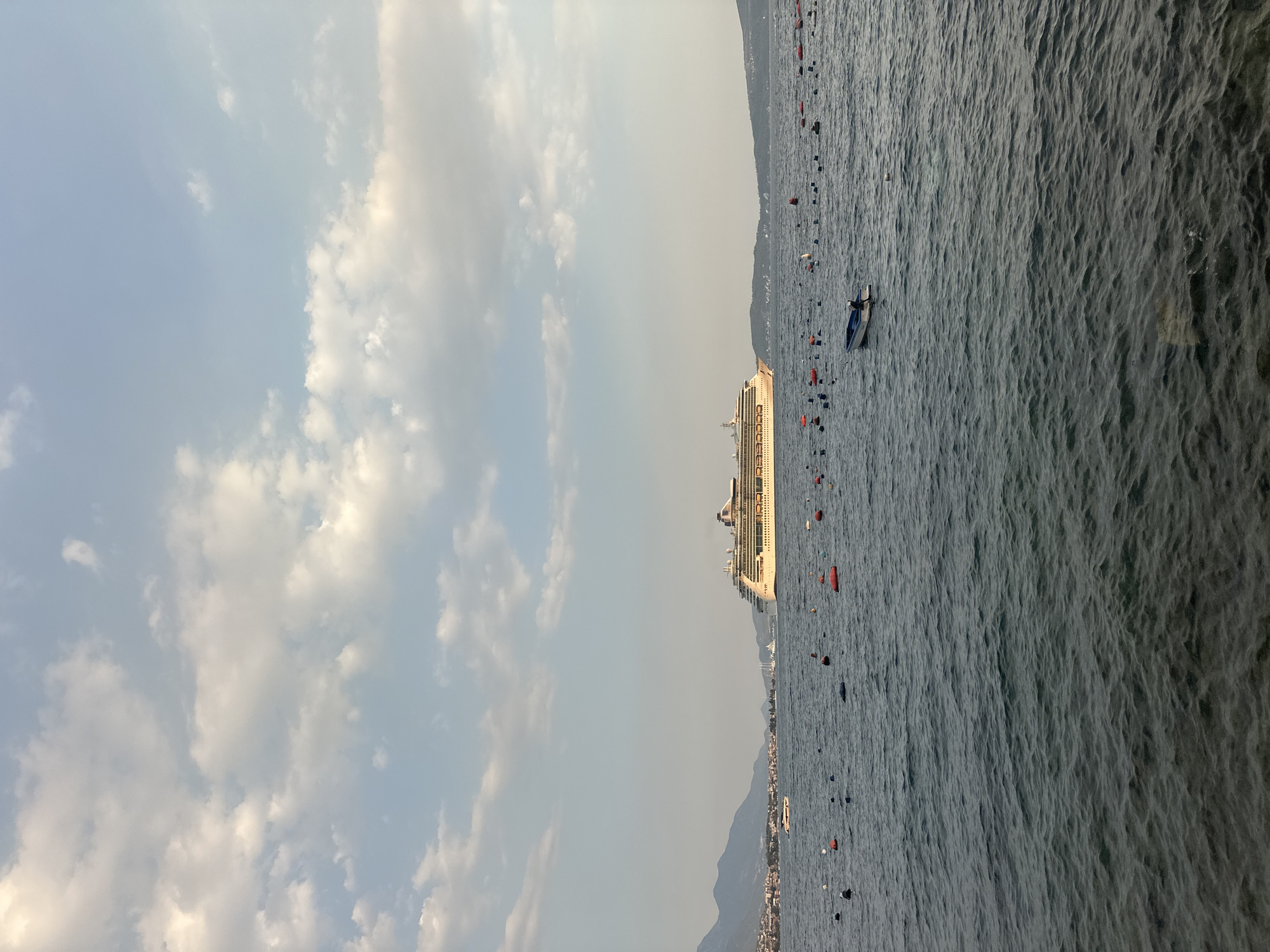 In Jošice, Montenegro, a large cruise ship is visible sailing on a calm, blue sea under a sky filled with scattered white clouds. The ship is white with a tan superstructure and appears to be moving towards the right side of the frame. In the foreground, numerous buoys of various colors, including red and blue, dot the water, suggesting an aquaculture or mooring area. A small, dark-colored boat with a person inside is positioned to the right of the center of the frame, amidst the buoys. In the distance, a coastline with buildings and mountains can be seen. The lighting suggests it is either early morning or late afternoon.