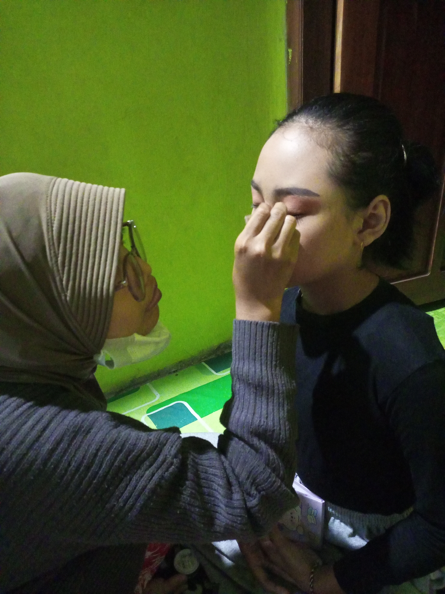 In Nanggulan, Indonesia, two individuals are captured in a dimly lit indoor setting. The background features a vibrant green wall, contrasting with a wooden door and a tiled floor in shades of green and white. On the left, a person wearing a grey hijab, glasses, and a mask is applying makeup to another individual on the right. This person is wearing a black long-sleeved shirt. The person receiving makeup has their eyes closed and their fingers are touching their eyelid. A small, pink, rectangular object with cartoon characters is visible on the floor in front of them. The overall scene suggests a personal grooming activity.