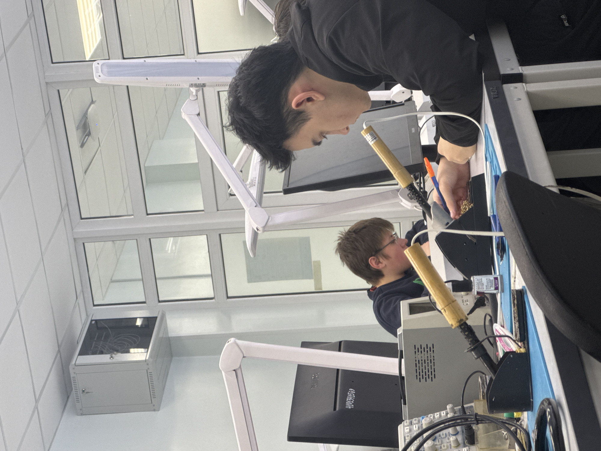This image depicts an indoor scene in an electronics laboratory or workshop, likely located in Perm, Russia, with two individuals engaged in precision work. The original photo is oriented vertically, rotated 90 degrees counter-clockwise.
In the foreground, a young man with short, dark hair, dressed in a black hooded sweatshirt, is leaning over a workbench, intensely focused on a task. He holds a tool, possibly a soldering iron with a distinct yellow handle, with his right hand, working on electronic components. His concentrated expression suggests meticulous attention to detail. A "UFC" logo is partially visible on his sleeve.
Further back and to the left, another young person with short, light brown hair and glasses is similarly engrossed in activity at a neighboring workstation, partially obscured by equipment. Both work areas are equipped with adjustable LED task lamps, illuminating their tasks. The workbenches host a variety of electronic equipment, including soldering iron holders, a grey power supply unit with visible vents and control knobs, and an assortment of cables. A "PHILIPS" brand monitor, displaying "4101344569" on its back, is situated behind the second individual. A pen with an orange cap is also visible on the main subject's workbench.
The room features large, white-framed windows reflecting the interior. The walls are white and paneled. A grey, rack-mount enclosure filled with cables is placed beneath the windows. The overall setting appears to be a modern educational or professional environment, indicative of electronics assembly, repair, or experimentation.