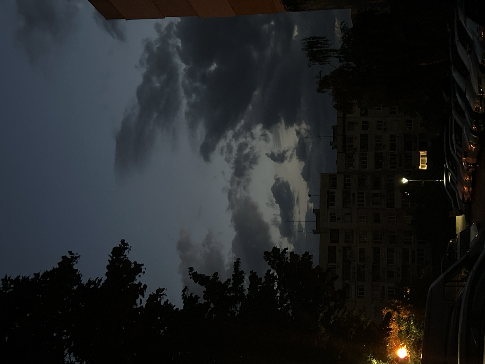 A nocturnal scene unfolds with a large apartment building dominating the mid-ground under a twilight sky filled with dark, heavy clouds. The building, with its rows of windows, is dimly lit, with a few windows showing interior light. In the foreground, a parking lot is filled with parked cars, their headlights or taillights casting soft glows. To the left, a silhouetted tree with dense foliage frames the view, and a streetlamp illuminates the branches. To the right, the side of another building, a light orange color, is partially visible. The overall atmosphere is somber and quiet.