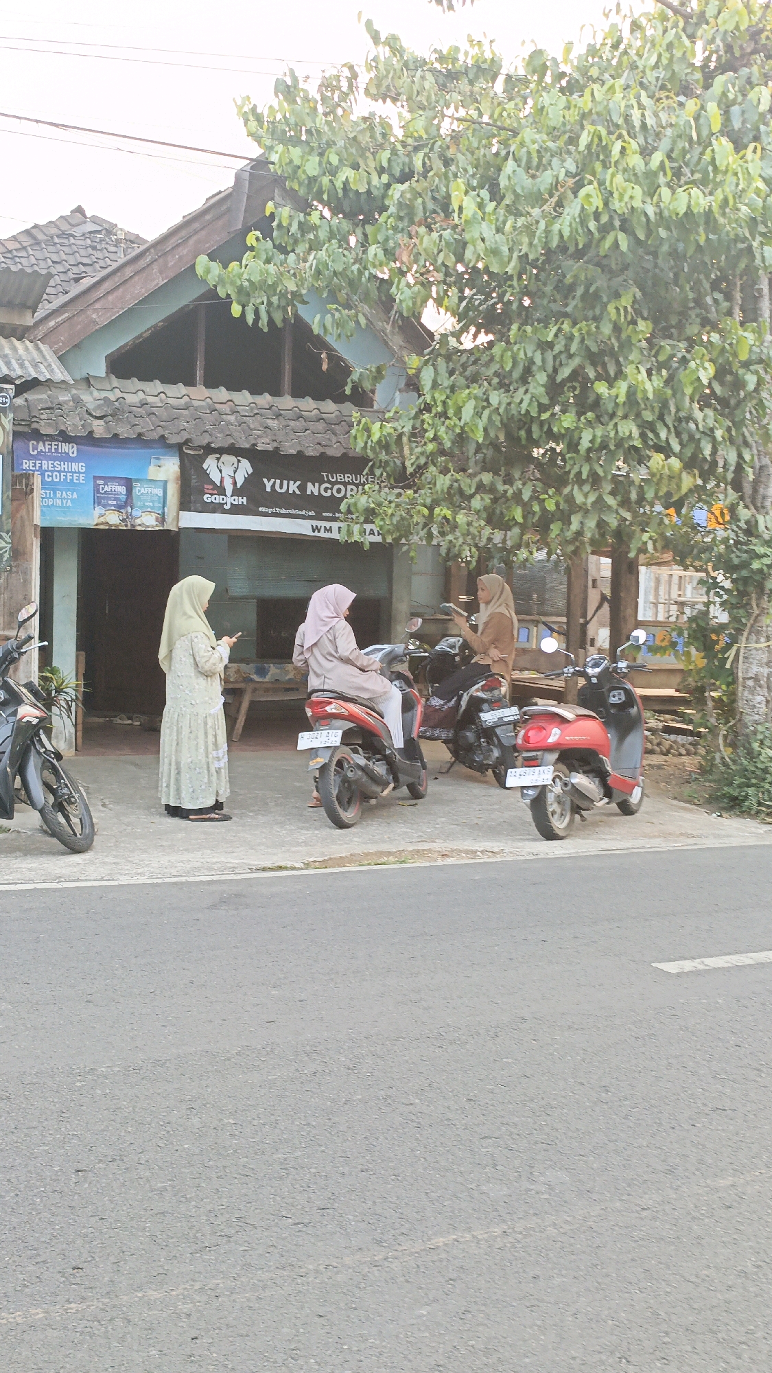 In Salatiga, Indonesia, a street scene unfolds in front of a shop with a sign reading "YUK NGOPIS". Three women are present. One woman, wearing a light green hijab and dress, stands on the left, looking at her phone. Two other women are seated on motorbikes. The woman on the red motorbike in the center wears a light pink hijab and dress, also looking at her phone. The woman on the black motorbike on the right wears a brown hijab and top, also engaged with her phone. Two motorbikes are parked in front of the shop, and a third, red motorbike is parked to the right. The shop appears to be a cafe or convenience store, indicated by posters for "CAFFINO REFRESHING COFFEE". A large tree with dense green foliage overhangs the scene. The ground is paved with asphalt, and a sidewalk is visible. The sky is overcast.