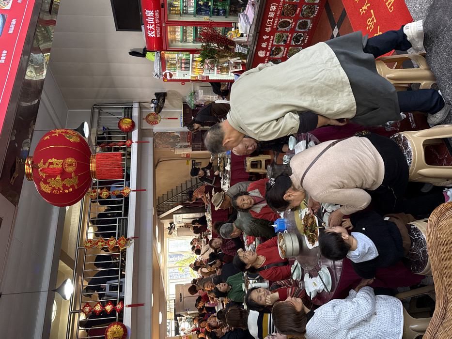 The image depicts the interior of a bustling multi-level Chinese restaurant in Yuzhong District, China. A large red Chinese lantern with gold characters and red tassels hangs prominently from the ceiling, surrounded by smaller red decorative elements and banners, indicating a festive or traditional ambiance.
The ground floor features numerous round tables, predominantly occupied by groups of individuals, mostly adults and elderly people, seated on plastic stools. Several tables have central lazy Susans loaded with various food dishes, bowls, and chopsticks. Interactions include people eating, conversing, and a seated elderly woman assisting a young child with food. An elderly man in a light-colored shirt and dark apron stands near a table on the right, facing away from the camera.
In the background on the right, two red "Nongfu Spring" refrigerators are visible, stocked with beverages. Above them, text reads "KUNGFU SPRING". Below the refrigerators, a menu board displays various dishes with images and Chinese text. A red floor mat with gold Chinese characters ("欢迎光临" - "Welcome") is positioned near the entrance.
An open staircase with black metal railings leads to a second floor, where more individuals are visible seated at tables, partially obscured by the railing. The restaurant appears well-lit by overhead fixtures and natural light from a window visible in the background on the left. Staff members are identifiable in the background, attending to tables and working near what appears to be a kitchen or serving area.