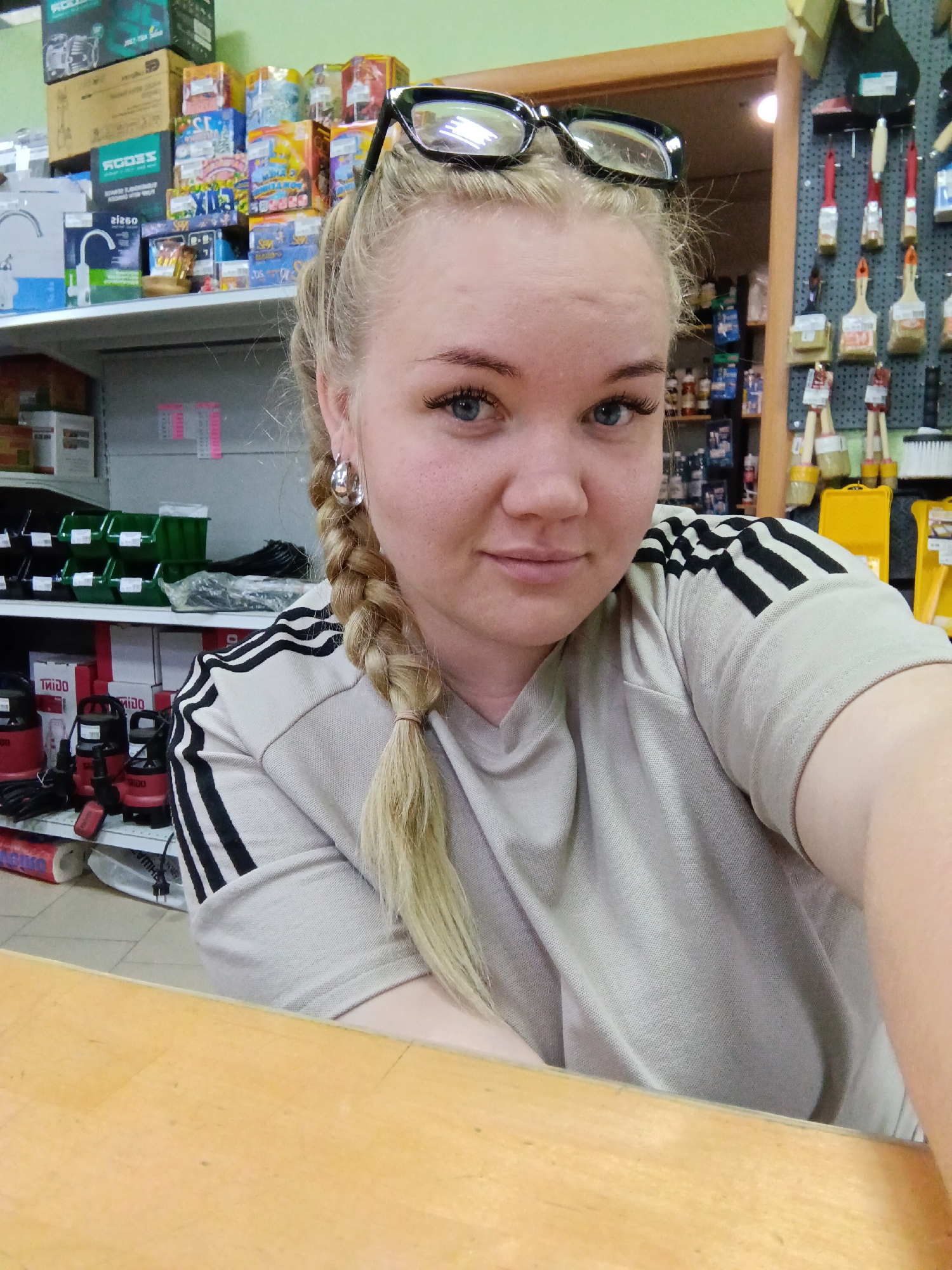 A blonde woman with a braid over her left shoulder is shown from the chest up, looking at the camera. She is wearing a tan-colored shirt with three black stripes on the shoulders. Glasses rest on top of her head. She has light skin, blue eyes, and is wearing silver hoop earrings. The background contains shelves filled with various items, including boxes, tools, and supplies commonly found in a hardware store. The location is not specified in the image. The city Фоки, Russia is not visibly confirmed by items in the background. The woman appears to be standing behind a light brown wooden counter.