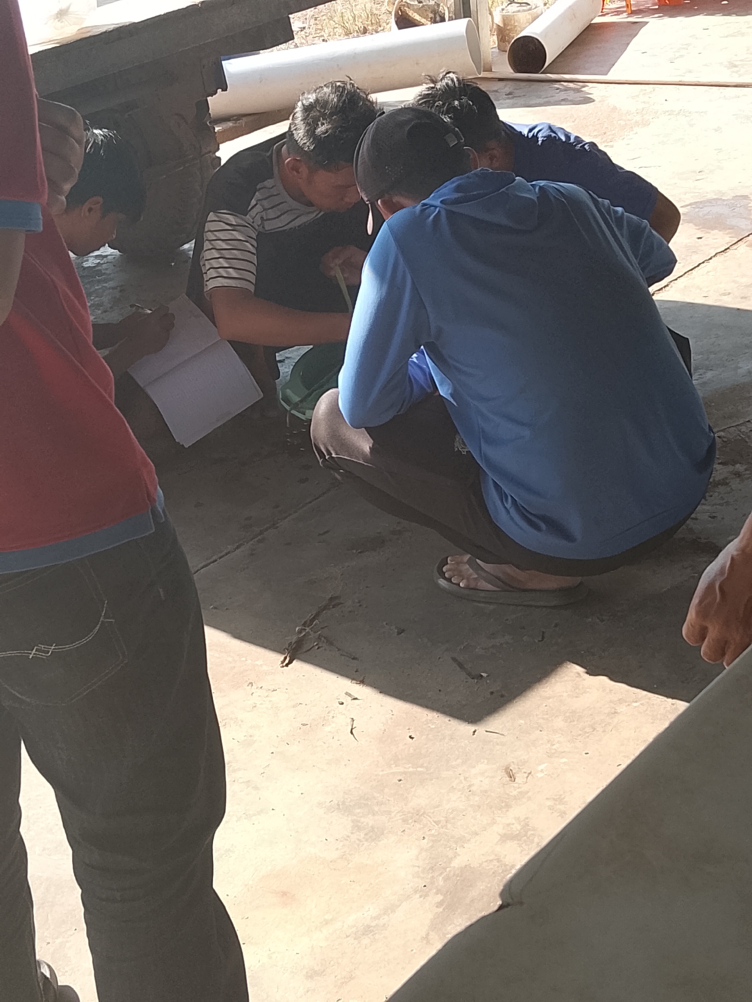 In Bukit Ketok, Indonesia, four individuals are observed on a sunlit, concrete ground. One person, wearing a red shirt and blue-trimmed collar, stands to the left, partially visible, facing away from the camera. To their right, a young individual in a red shirt is crouched, writing in an open notebook with a pen. Three other individuals are in the midground, also crouched or kneeling. One person, wearing a black and white striped shirt, is looking down and appears to be interacting with something in a green container. Beside them, a person wearing a blue hoodie and black pants, with a black cap, is also looking down, their focus directed towards the ground. Another individual, wearing a blue shirt, is partially visible behind the person in the blue hoodie. A large grey object, possibly part of a vehicle or structure, is present in the background to the left. White pipes are visible in the further background. The scene is illuminated by natural sunlight, casting shadows on the ground.