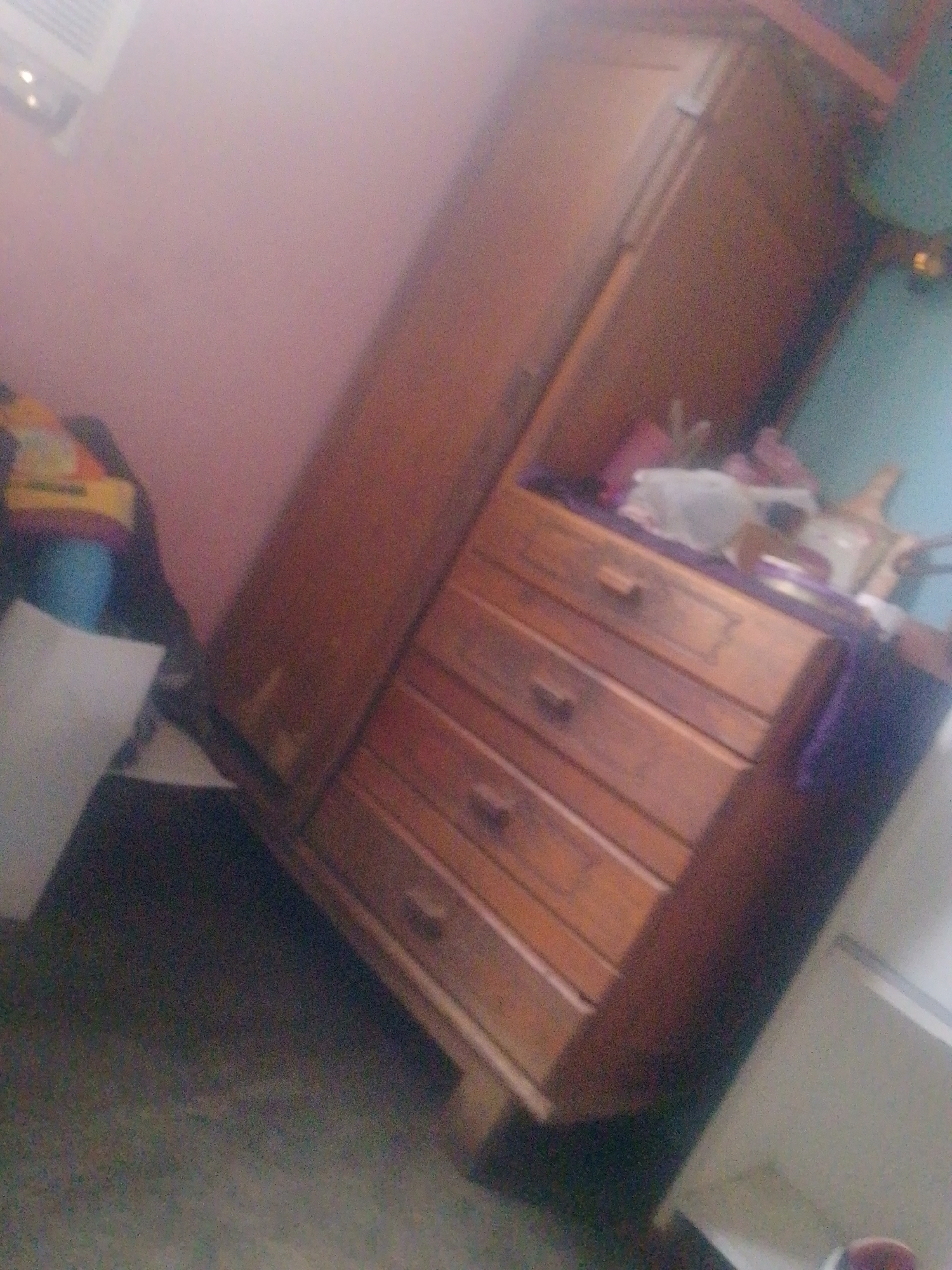 The image shows a wooden wardrobe with drawers, leaning slightly to the left. The wardrobe is placed against a wall that is pink on one side and light blue on the other. There is a white trash can to the left of the wardrobe. The lighting is dim. The exact location in Juan Griego, Venezuela, cannot be determined from the image.