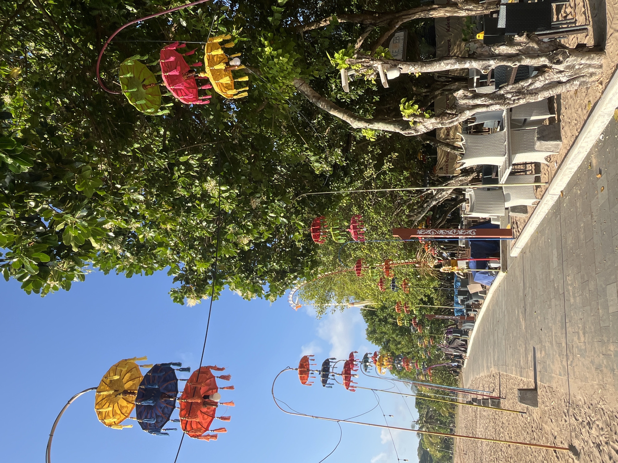 The image depicts a pathway lined with colorful, decorative umbrellas hanging from poles along a sandy beach in Kuta Selatan, Indonesia. The pathway is paved with grey tiles and runs adjacent to a row of outdoor seating for a restaurant, complete with tables and chairs. Lush green trees with dense foliage overhang the seating area, casting shadows on the ground. In the background, a Ferris wheel is visible above the trees. The sky is clear and blue, indicating a sunny day. Several people are seen walking on the pathway, some in the distance, and there are no interactions between them. The scene appears to be a tranquil beachside setting, possibly in the morning or late afternoon, given the quality of light.