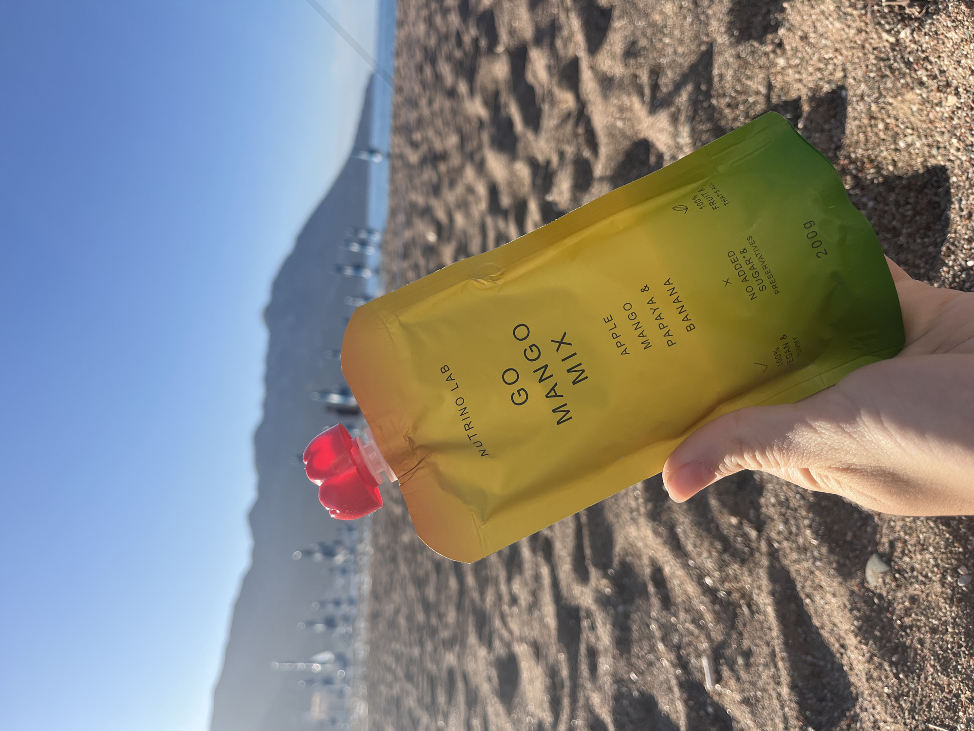 The image depicts a yellow and green package of "Go Mango Mix" held in a hand on a beach. The package specifies ingredients such as apple, mango, papaya, and banana. The label also indicates "No Added Sugar & Preservatives", and that it is "100% Vegan & Yummy". The packaging indicates it is from "Nutrino Lab" and weighs "200g".
The background shows a beach, with the sea and the mountains of Budva, Montenegro visible in the distance. The foreground is filled with sand and some blurry details. A mountain and a town can be seen to the left of the image. The sky is blue and clear.