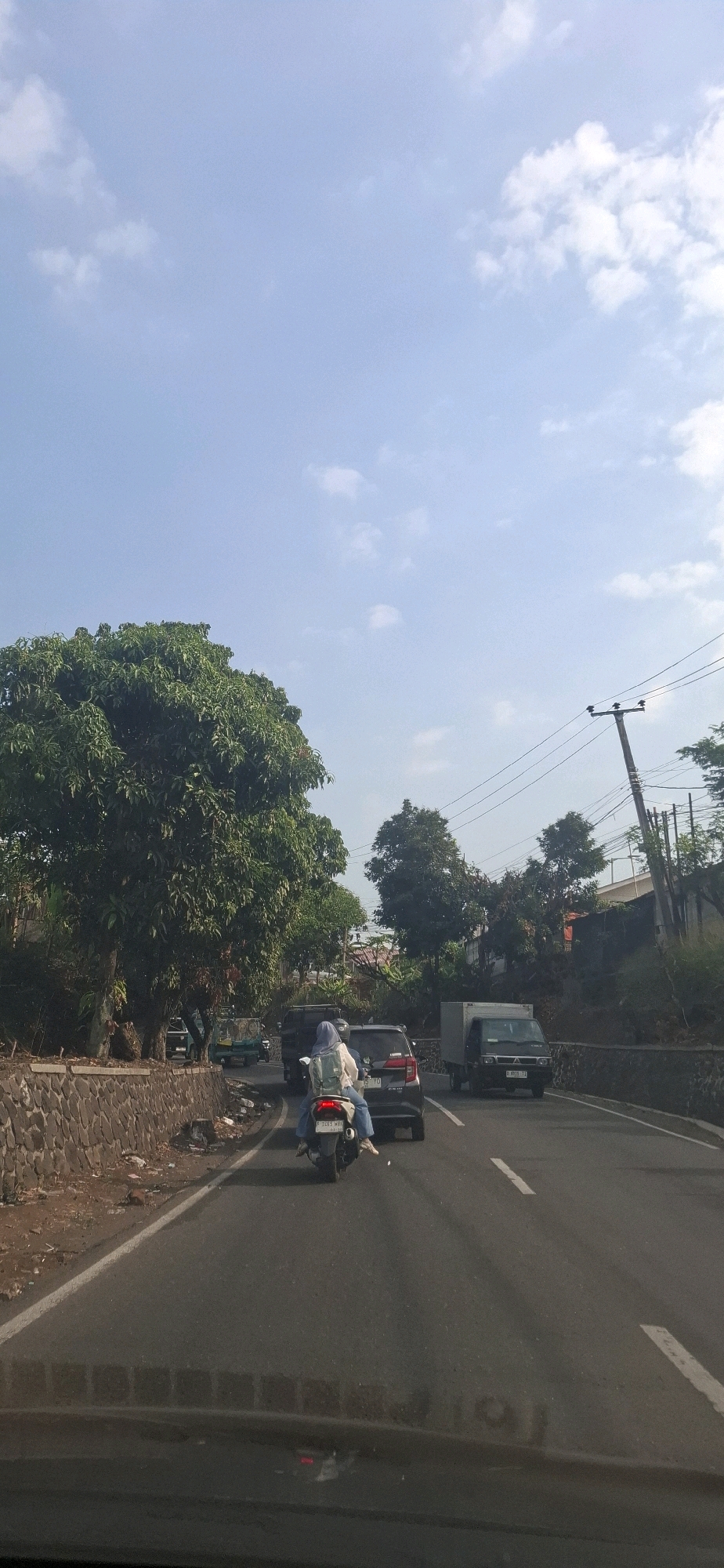 The image depicts a view from a vehicle on a road in Cilaku, Indonesia. The sky is clear with some scattered white clouds. On the left side of the road, there is a large, leafy green tree and a stone wall. Several vehicles are on the road, including a motorcycle with a rider wearing a light blue hijab and a backpack, a black SUV, and a light-colored truck. A utility pole with wires is visible on the right side of the road, and further in the distance, more trees and buildings can be seen. The road appears to be a two-lane highway with a white line dividing the lanes. The overall scene suggests a daytime view of traffic on a road in a somewhat rural or suburban area.