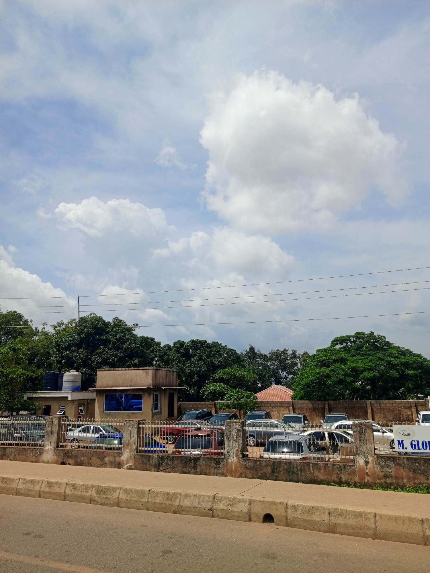 The image depicts a street scene in Kabong, Nigeria, with a clear blue sky filled with fluffy white clouds. A road runs in the foreground, bordered by a concrete barrier. Behind the barrier, there is a fenced area containing several parked cars. A building with a light brown facade and blue-tinted windows is visible behind the fence. Two water tanks are mounted on the roof of the building. Lush green trees and vegetation form a backdrop to the scene, with power lines strung across the upper portion of the image. The atmosphere suggests a sunny day in a somewhat developed urban area. There are no people visible in the image.