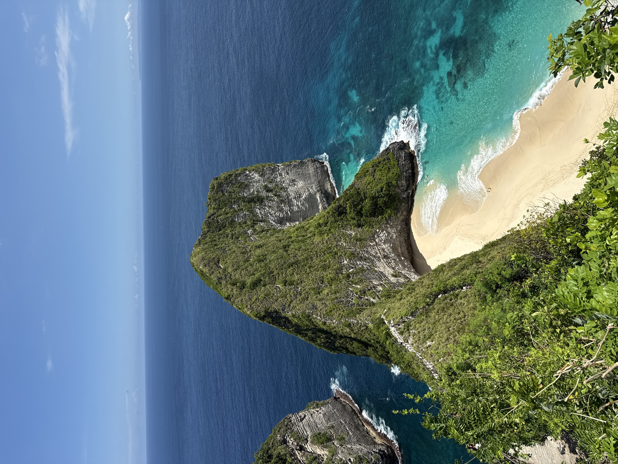 The image depicts an aerial view of Kelingking Beach, located in Bunga Mekar, Nusa Penida, Indonesia. A large, T-shaped headland covered in dense green vegetation extends into the turquoise and deep blue ocean. The headland features a steep, rugged cliff face with visible rock strata. A sandy beach, edged by white foamy waves, nestles at the base of the headland. A walking path is visible along the ridge leading to the beach, and there are 2 people visible in the sand. The sky is clear blue with some wispy clouds.