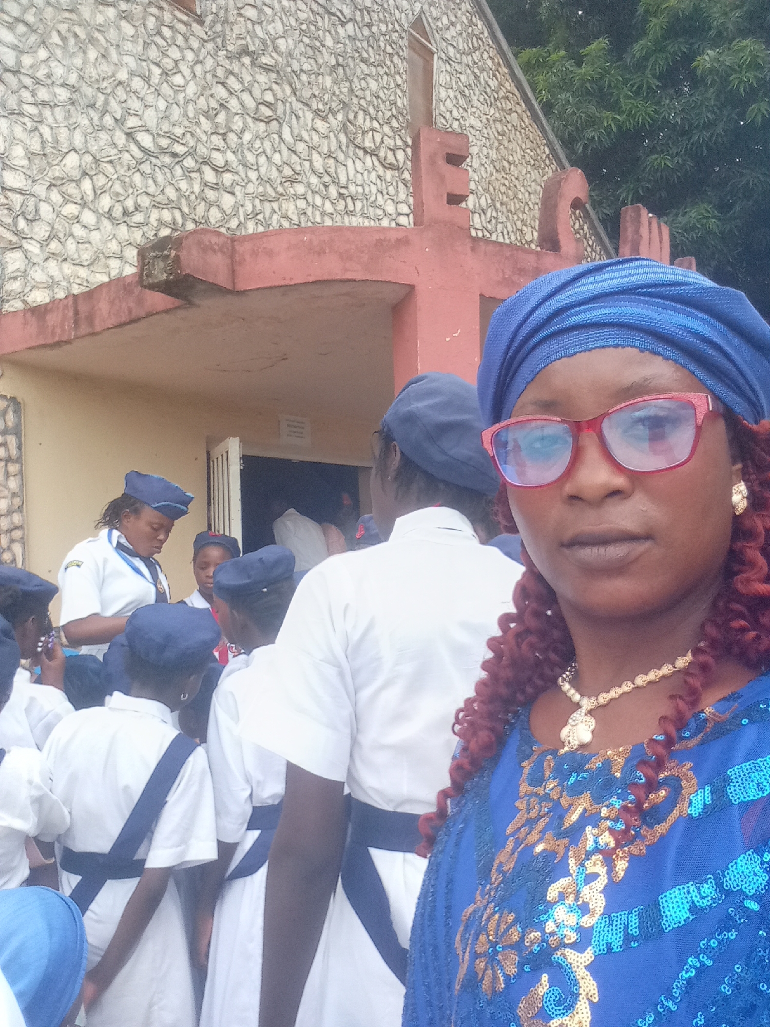 The image captures a group of people in front of a building with a stone exterior and a prominent cross on top. The location is in an unknown city, within an unknown state/province. The group seems to consist of young women and at least one adult female dressed in blue and wearing red glasses. The young women appear to be in a uniform of white shirts with dark blue straps and dark blue caps. The adult woman in the foreground is wearing a sequined blue dress and a blue headwrap. Some individuals are entering the building, while others are standing outside.