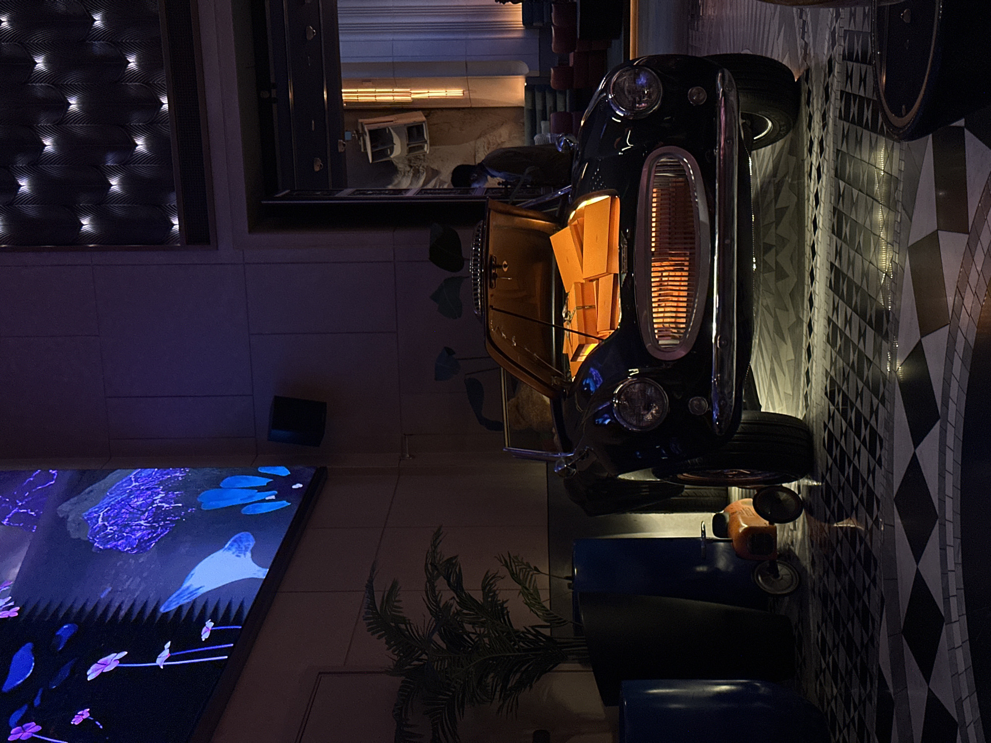 The image captures a dimly lit indoor space, rich with decorative elements, suggesting an upscale lounge, restaurant, or hotel lobby. A prominent black, vintage-style miniature car is positioned on its side, dominating the upper-right portion of the frame. Its hood is open, emitting a striking warm orange glow from within, illuminating its interior. The car features classic design elements, including round headlights and chrome accents.
To the right of the car, a floor boasts a bold black and white geometric tiled pattern, adding a visually engaging texture to the space. Below the car, a dark blue seating area is visible, contributing to the lounge-like atmosphere. Near the car's rear, an orange, retro-styled object, possibly a small scooter or a decorative gas pump, adds another vintage touch.
On the left side of the frame, a large digital screen displays vibrant, abstract patterns in shades of blue and purple, resembling fluid organic shapes or ethereal flora. Above this screen, a dark wall panel is adorned with a textured pattern and numerous small, bright white lights, creating a soft, ambient illumination. In the upper-left background, a reflection or an open area reveals a glimpse of an interior with a counter or desk, behind which a person can be vaguely discerned. A faux palm plant rests on the floor in the lower-middle section.
The overall ambiance is sophisticated and modern, yet incorporates nostalgic elements. The subdued lighting indicates an evening or nighttime setting. No visible text is present in the image. This type of elaborate and stylish interior design aligns with establishments found in Dubai, United Arab Emirates.