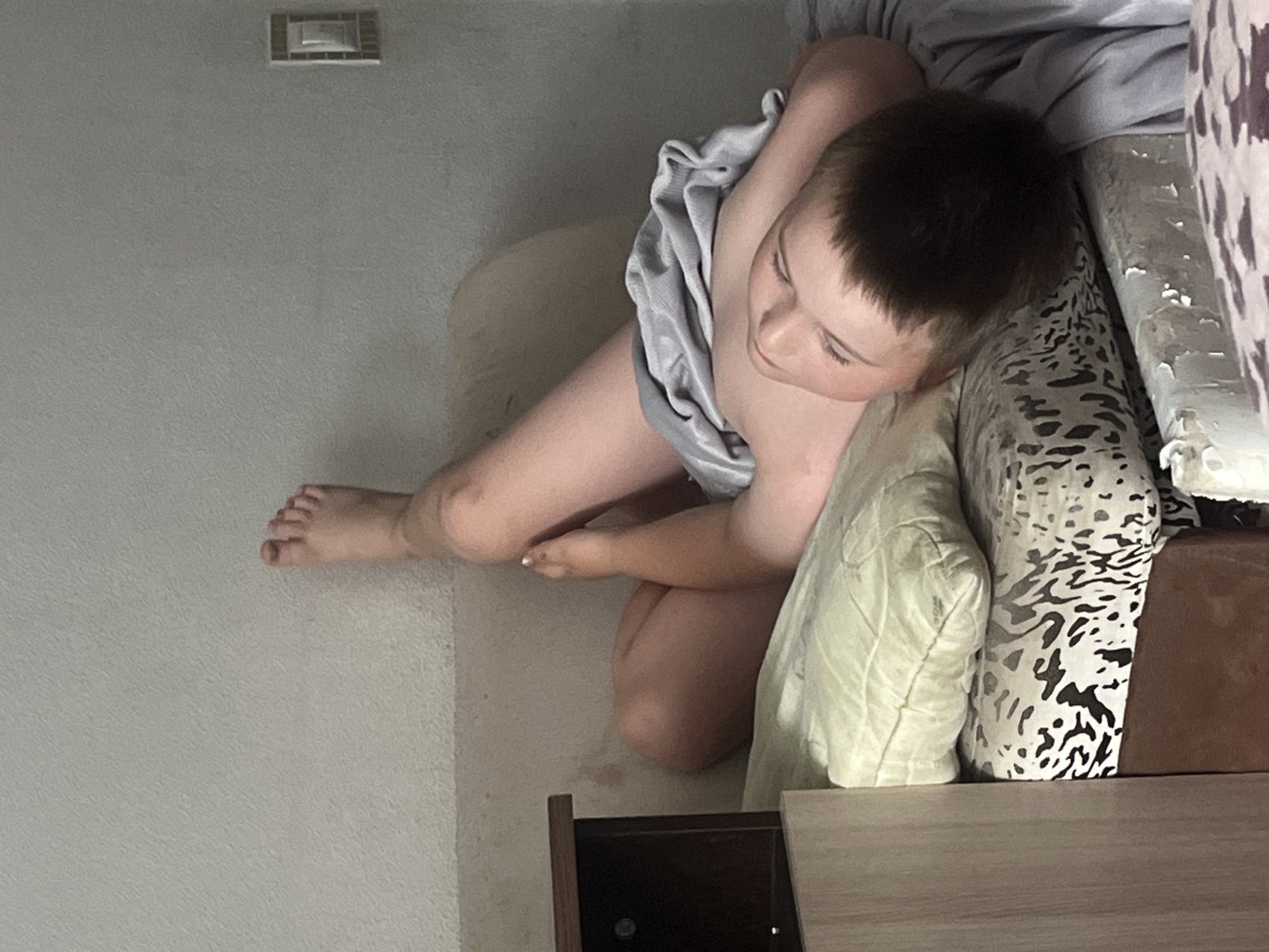 The image shows a young boy lying on a bed, partially covered by a grey blanket. His head is resting on a pillow, and his eyes are closed. One of his legs is extended upwards, with his foot visible against a light-colored wall. To the left of the bed, there is a dark-colored nightstand. The bed has a patterned sheet and another pillow visible. The background consists of a plain, light-colored wall with a light switch on the right side. The overall impression is that of a bedroom scene. The provided information states this image is located in Chelyabinsk, Russia.