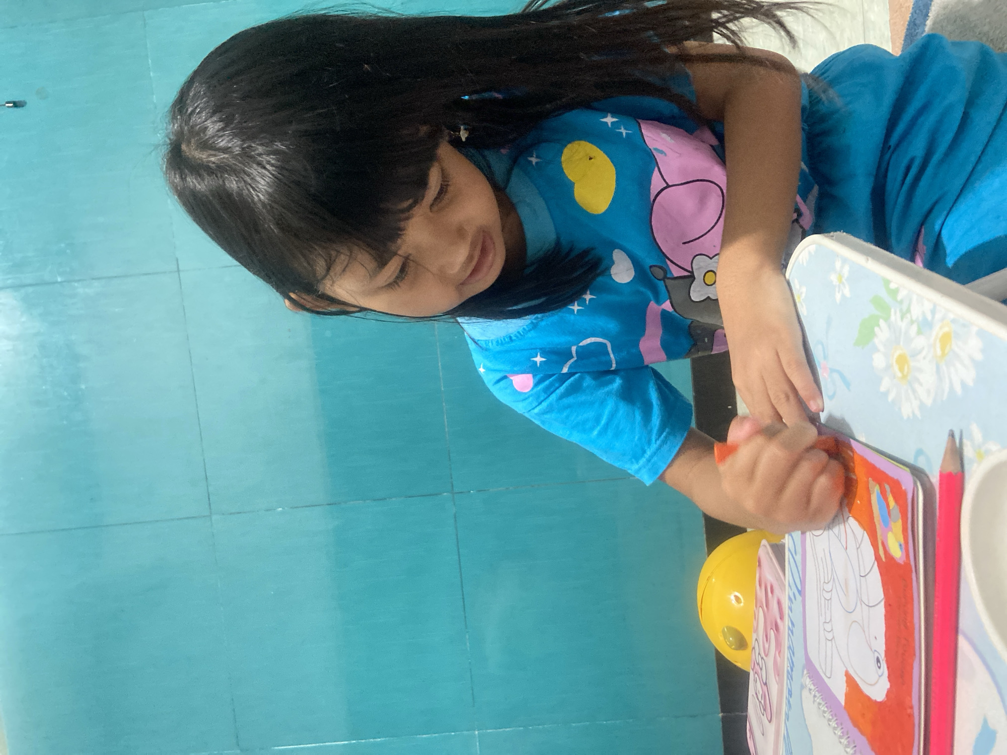 A young, dark-haired child in a blue, short-sleeved shirt is sitting at a small table, absorbed in coloring a picture in a book. The table is light blue and decorated with white flowers. The coloring book is brightly colored with a picture of a cartoon character on the page she is working on. The child is holding an orange crayon in her right hand and appears to be pressing down firmly as she colors. To the right of the coloring book is a pink pencil and a small white container. A yellow toy is sitting near the table's edge. The floor is turquoise tile with visible grout lines. It can be inferred that the location of the image is inside a home in Gelam, Indonesia.