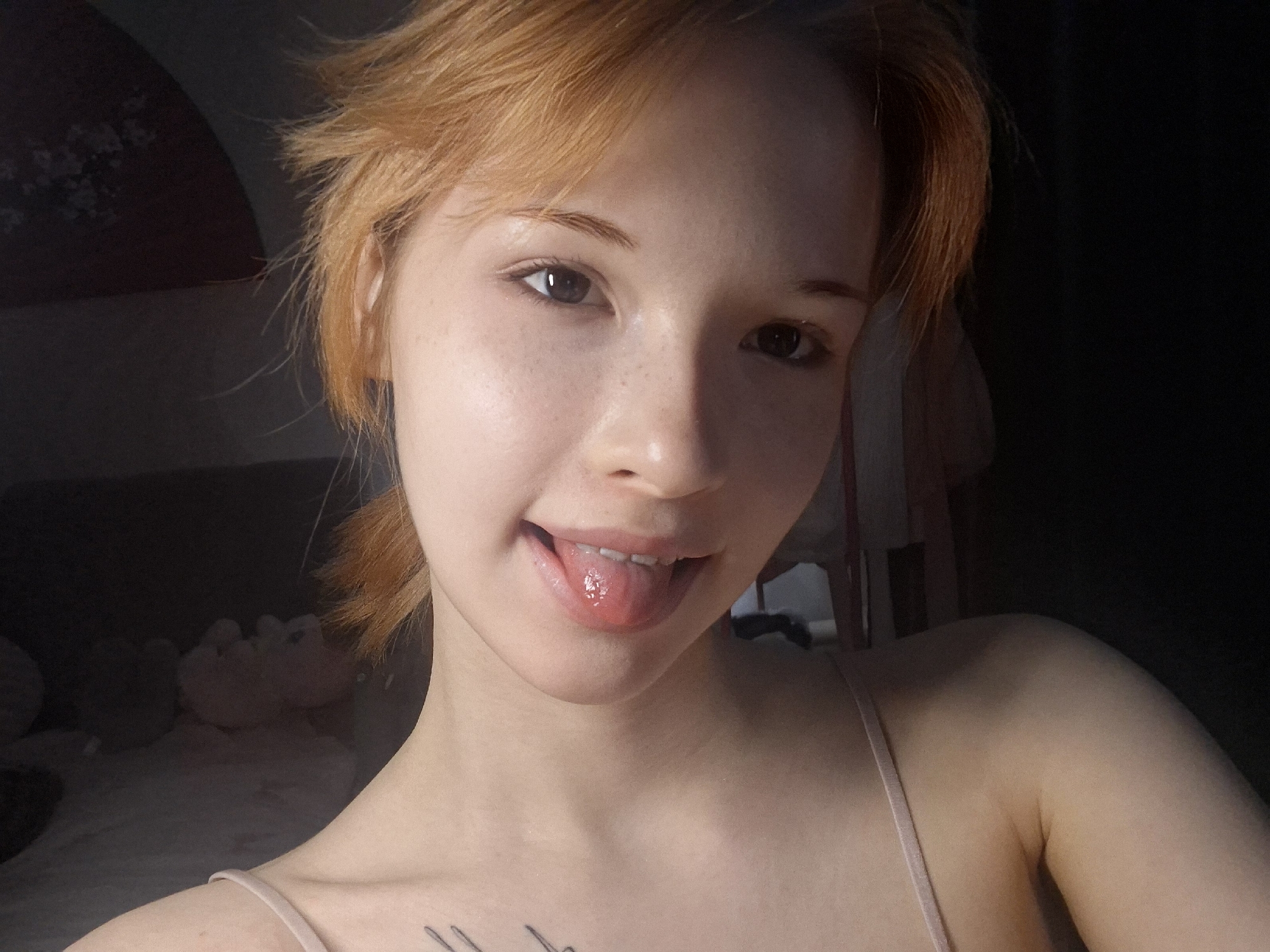 A close-up selfie captures a young individual, likely female, with fair skin, dark eyes, and tousled, reddish-blonde hair. She looks directly at the camera with a playful expression, sticking out her wet and glistening tongue while a faint smile plays on her lips. She is wearing a light-colored, possibly nude or light pink, spaghetti strap top. A linear tattoo design is partially visible on her left clavicle/shoulder area. Her face is well-lit, highlighting her clear skin and a few faint freckles. The background is very dark and indistinct, suggesting an indoor setting with minimal ambient light. Vague, lighter shapes, possibly plush toys or furniture, are dimly discernible behind her left shoulder. The overall mood is casual and cheerful. This scene is set in Chaikovsky, Russia.