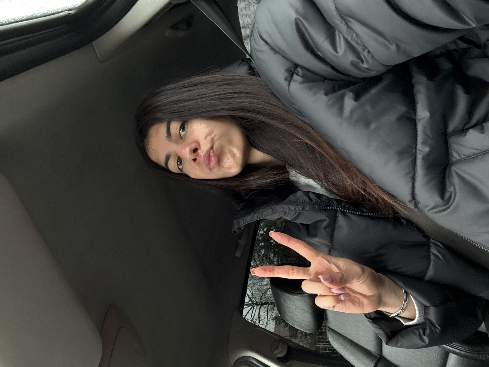 The image displays a young female seated within the interior of a vehicle, located in Sofia, Bulgaria. The subject has long dark brown hair and is looking directly at the camera with pursed lips. Her left hand is raised, forming a "peace" sign with her index and middle fingers, which have light pink nail polish. A silver-colored bracelet is visible on her left wrist. She is wearing a black puffer jacket over a light gray long-sleeved top. The vehicle's interior features dark-colored upholstery on the seat and headrest, with dark gray trim on the ceiling and pillars. A seatbelt is visible across the subject's chest. Through the car's left side window, bare deciduous trees are visible against an overcast sky, indicating an outdoor, non-summer environment.