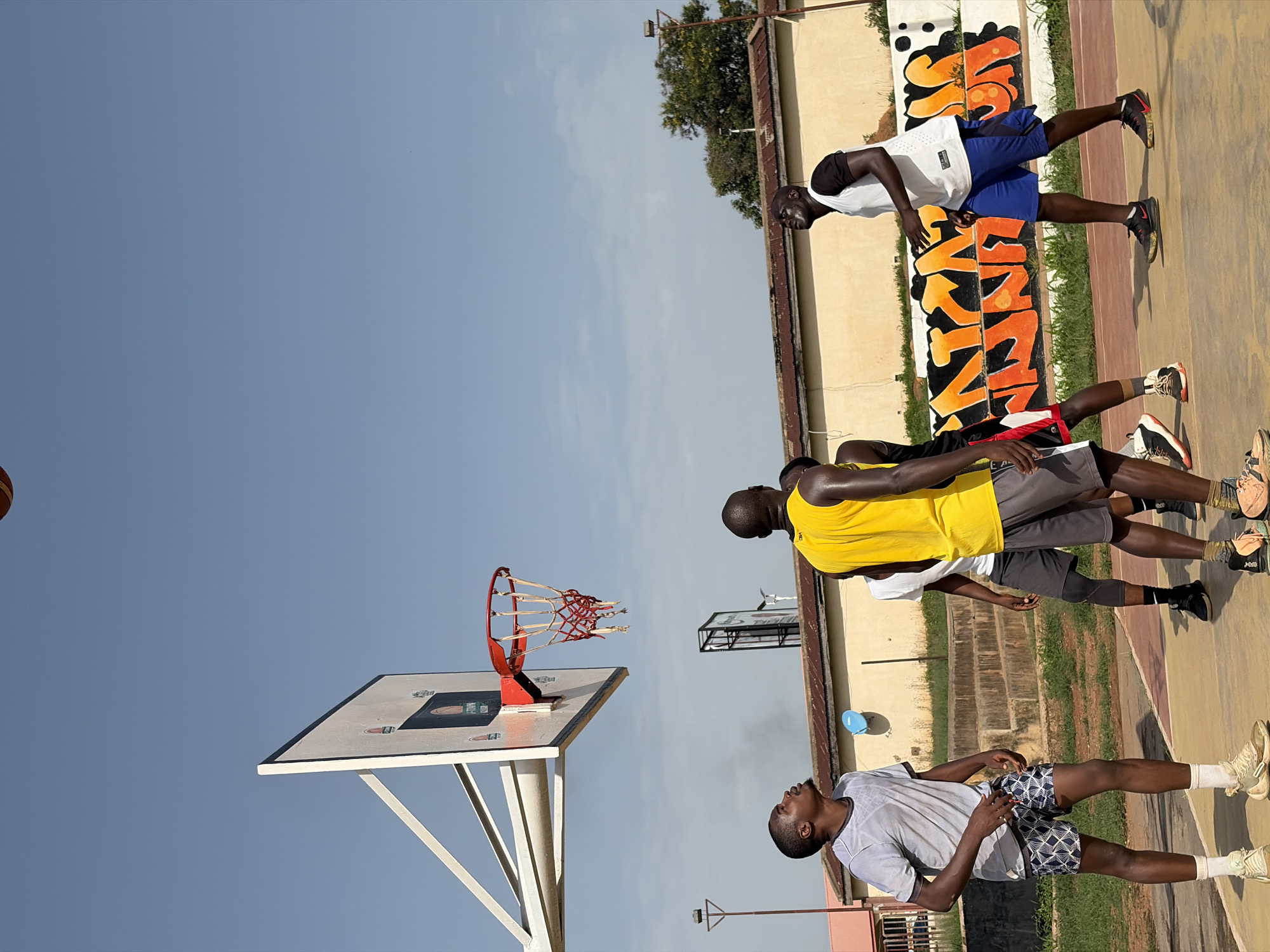 The image depicts a group of men playing basketball outdoors. They are on a court with a basketball hoop. One of the men is looking up towards the basket. Another man is wearing a yellow tank top. The location is in Jos, Nigeria. There is a building with graffiti on the side behind the basketball court. The sky is mostly clear blue.