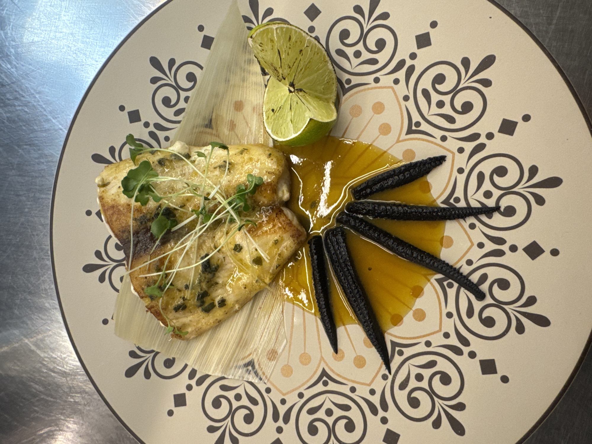 A beautifully plated dish of grilled fish rests on a decorative plate, accented by a lime wedge and a garnish of microgreens and what appear to be grilled baby corn. The fish, a pale white fillet, is golden brown and slightly charred on top, seasoned with visible herbs and drizzled with a glistening sauce. The plate itself features an intricate, mandala-like pattern with dark brown and orange details on a cream background. The overall scene is captured from a top-down perspective, likely within a restaurant kitchen or dining area, given the metallic surface visible in the background which suggests a countertop or table.
