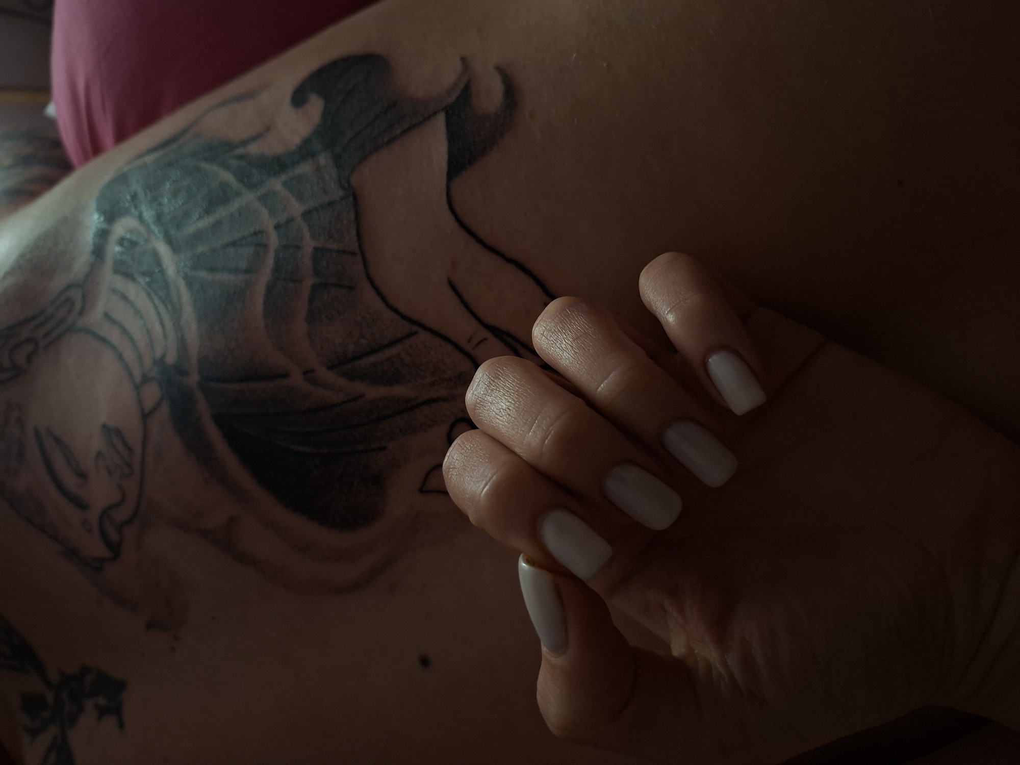 A person's hand with white manicured nails rests on a tattooed torso. The tattoo is large and black, covering a significant portion of the visible skin. The torso is partially visible, with a hint of pink clothing in the upper area. The lighting is dim, casting shadows across the scene. The location is unknown.