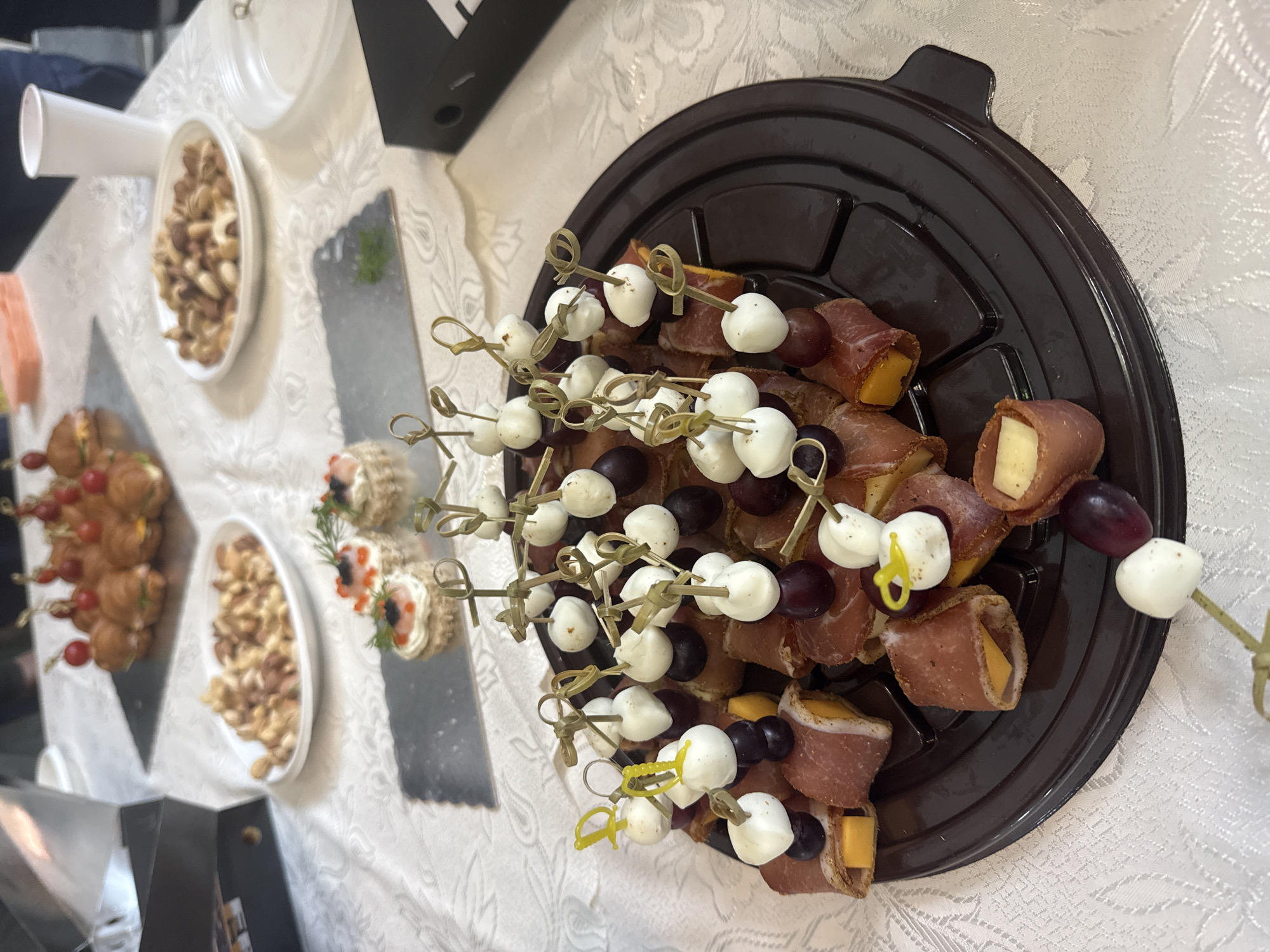 The image shows a table with several platters of food, likely at a catered event. There's a round, dark platter filled with skewers of mozzarella balls, grapes, and rolled prosciutto with cheese. Other food platters include small sandwiches on skewers, appetizers with red roe, and a white oval dish filled with nuts. The tablecloth is white with a subtle pattern. A white cup and what appears to be office supplies (black binder) are also visible on the table. The scene is set indoors, possibly in Sofia, Bulgaria, but there's nothing in the image to confirm the exact location. There are no people in the frame.