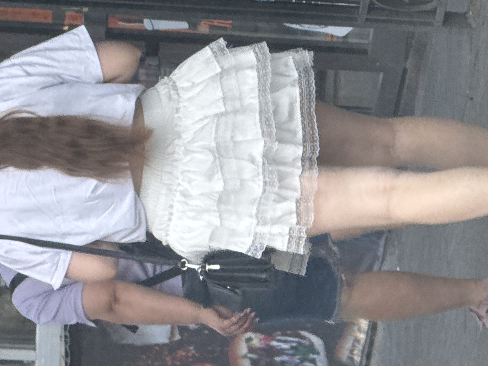 A person stands outdoors in a city, possibly Rostov-on-Don, Russia, with their back to the camera. They are wearing a white t-shirt, a dark skirt with ruffles and lace trim, and denim shorts. A black purse is slung across their shoulder. The person's hair is long and brown, falling down their back. In the background, a building with dark metal bars is visible, along with what appears to be a poster or artwork on a wall. The ground is paved.
