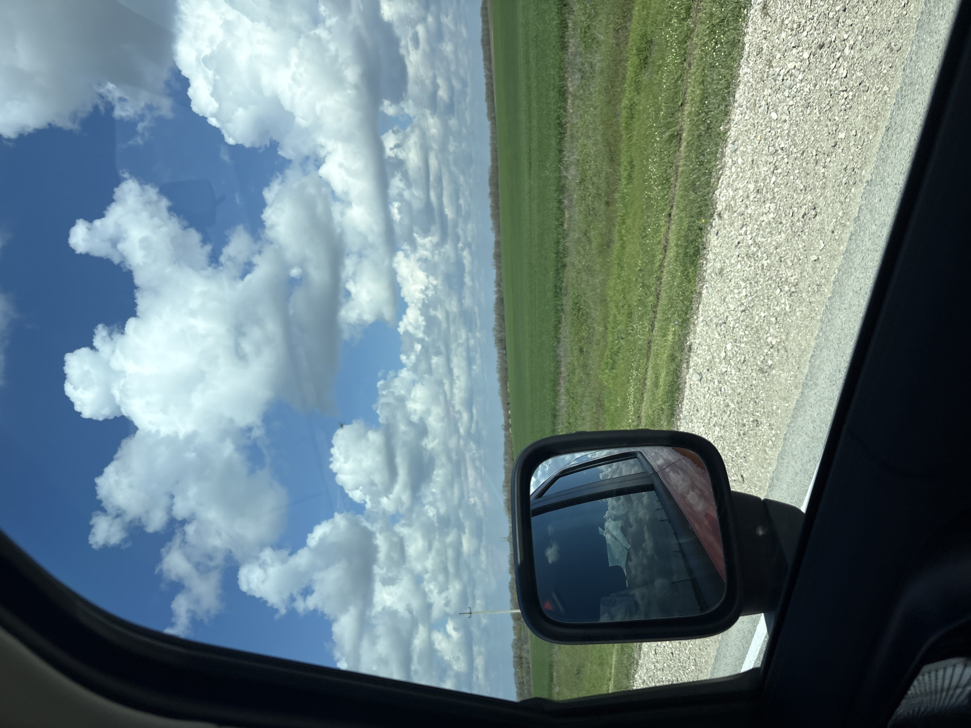 The image shows a view from inside a car. The scene is oriented sideways. A blue sky with fluffy white clouds dominates the view through the car window. A side mirror is visible, reflecting the sky. Outside, green fields extend along the roadside, adjacent to a gravel road. A power pole is seen in the distance. The exact location in Пожарское сельское поселение, Ukraine is indeterminable without further information.