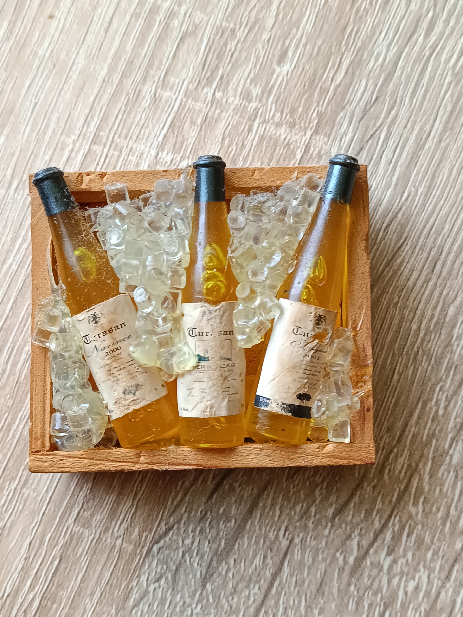 A miniature wooden crate containing three miniature amber-colored bottles filled with a yellow liquid is depicted. The bottles are surrounded by clear, translucent resin pieces shaped like ice cubes. Each bottle has a label with text that is partially obscured by the "ice." The crate and its contents are placed on a light-colored wooden surface with a wood grain pattern. The image is taken from a top-down perspective. There are no people or animals present. The location in the city of Tebras, Latvia is not discernible from the image.