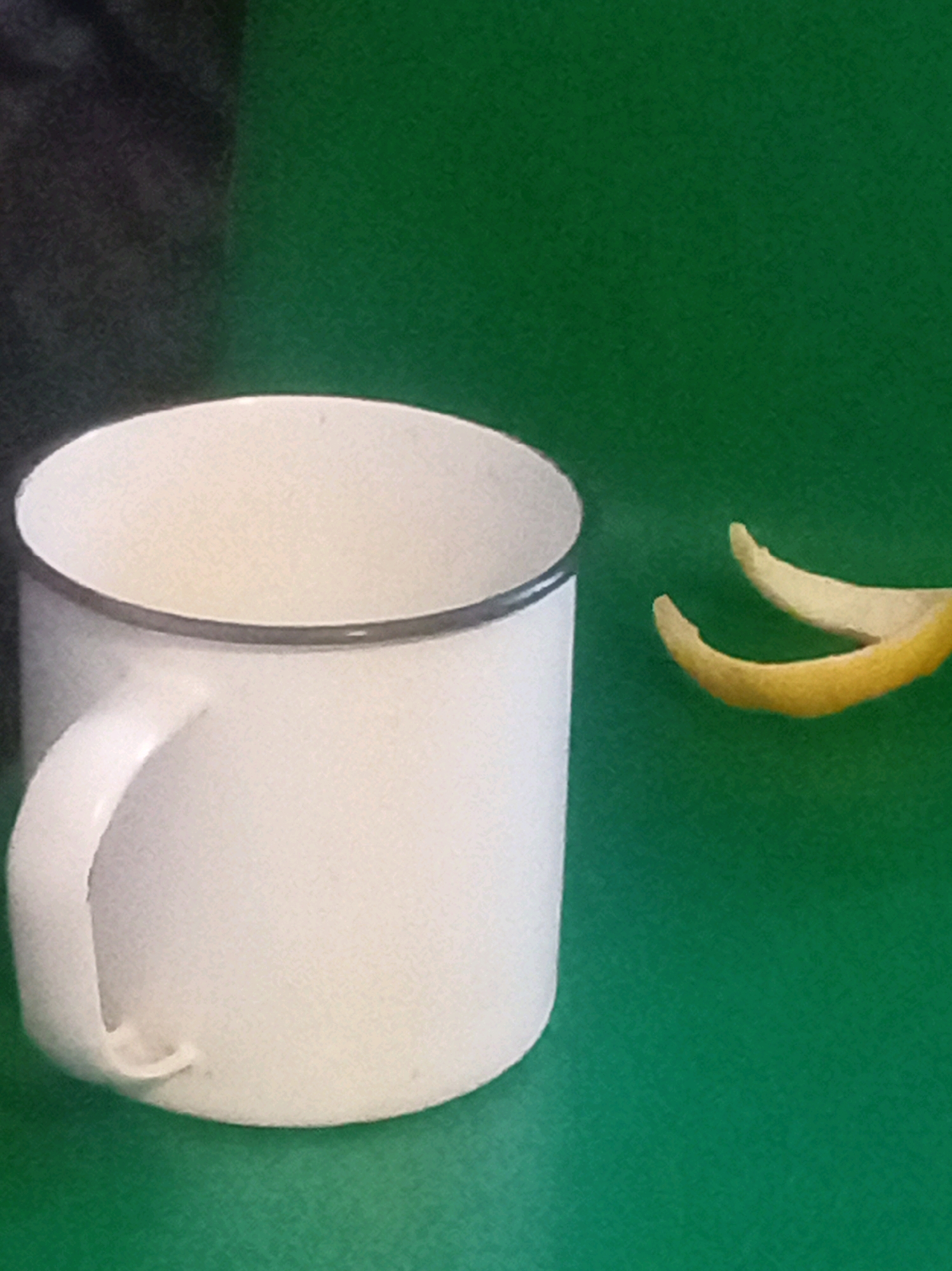 A white enamel mug with a dark rim sits on a green surface. To the right of the mug are two curved pieces of yellow peel, likely from a citrus fruit. The background is a solid green. No people or specific landmarks of Mhluzi, South Africa, are visible.