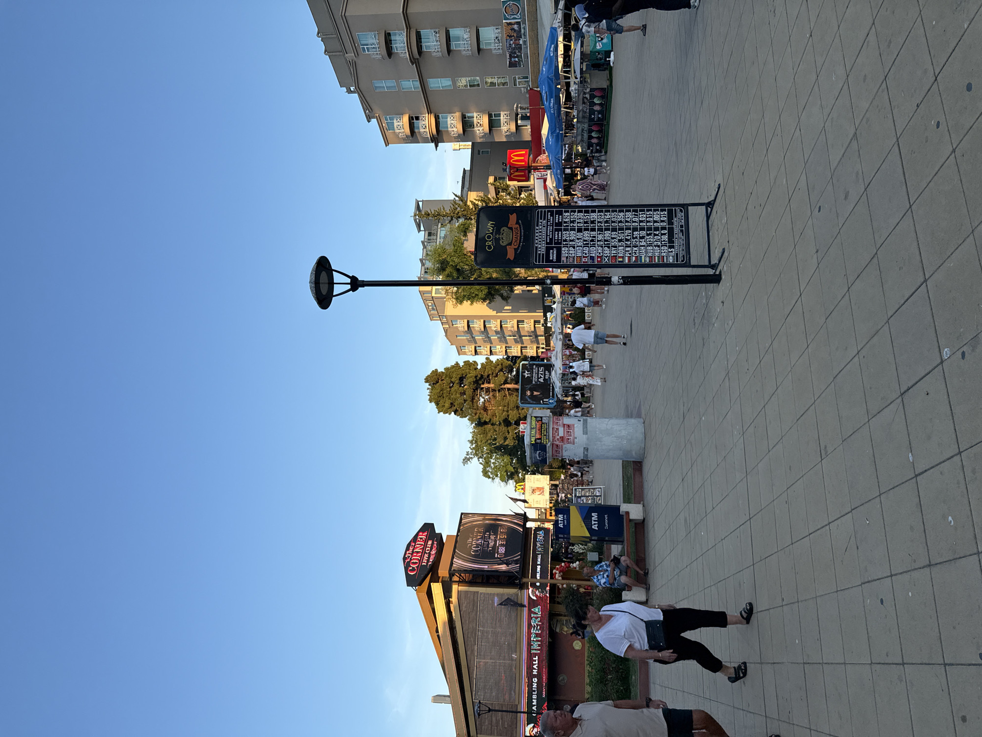 The image depicts a scene in the city of Nesebar, Bulgaria, specifically on a pedestrian walkway. The sky is clear and blue, indicating daytime. In the foreground, a paved walkway made of large square tiles leads towards the background. To the left, a man and a woman are walking. The woman is wearing a white top and black pants, and carries a dark bag over her shoulder.
Further into the scene, there are several buildings and businesses. A prominent building on the left has a sign that reads "The Corner Live Club" in illuminated red letters. Below this sign, another sign advertises "Gambling Hall IMPERIA". An ATM machine is visible to the right of this building.
In the center-right of the image, a tall black pole with a circular lamp at the top stands near a large black sign on an easel. This sign displays exchange rates for various currencies, with the title "CROWN CHANGE". Below the currency exchange board, a McDonald's restaurant with its distinctive golden arches is visible. Adjacent to McDonald's are several outdoor stalls with blue umbrellas, possibly selling food or drinks.
Several people are scattered throughout the scene, walking or standing, suggesting a public area with tourist activity. The overall environment appears to be a commercial or entertainment district, likely a popular spot for visitors in Nesebar. The buildings in the background are multi-story structures, some with balconies. Trees are visible behind some of the buildings, adding a touch of greenery to the urban landscape.