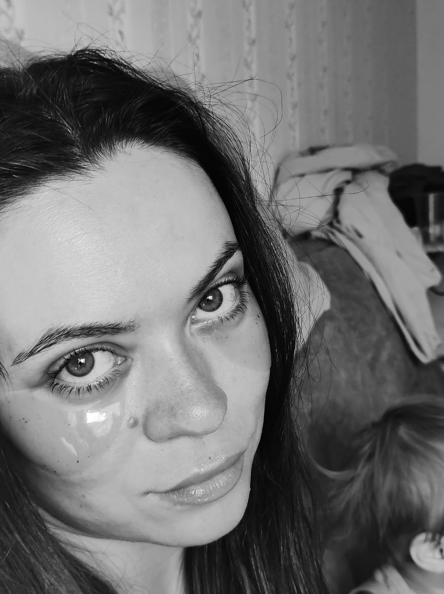A black and white close-up portrait of a woman with dark hair, facing the camera. She has eye masks on her face. The background is blurry and shows a room with what appears to be a pile of clothes and a glimpse of a small child. There is no evidence to suggest the location is Chaikovsky, Russia.