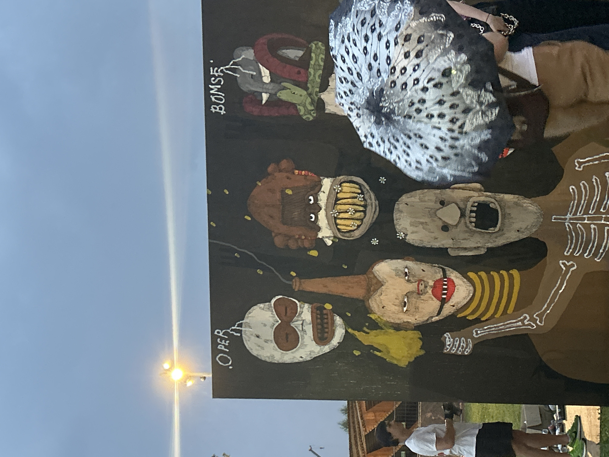 A group of people are gathered around a large, dark mural painted with various figures. The mural features several stylized faces with exaggerated features and bold colors. To the left, a person wearing a white t-shirt and black shorts stands in profile, observing the artwork. To the right, a person is partially visible holding an umbrella decorated with a peacock feather pattern. The background shows a cloudy sky and some distant structures, suggesting an outdoor setting in a city. The overall atmosphere appears casual and social, with people interacting with the art.