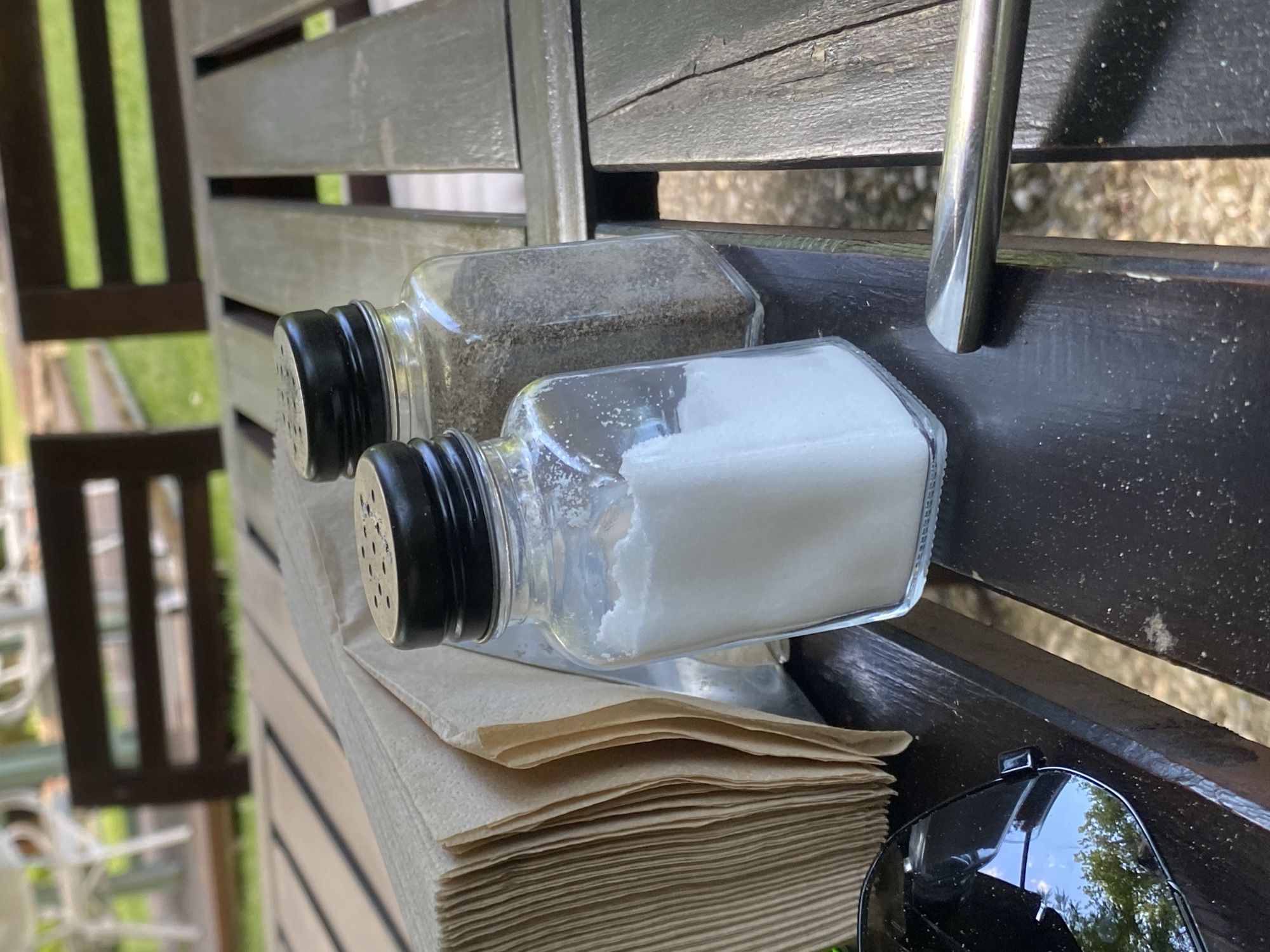 The image shows a close-up view of a wooden table, likely outdoors, with a stack of brown paper napkins to the left and two glass salt and pepper shakers. The shaker in the foreground is filled with white salt, and the one behind it contains black pepper. A pair of black sunglasses rests on the left side of the table, reflecting a green, leafy tree and blue sky. A portion of a metallic utensil, possibly a fork or spoon, is visible on the right side of the table. The background is blurred, showing out-of-focus outdoor seating and greenery, suggesting a dining or seating area. The scene appears to be set in Varna, Bulgaria, based on the provided context.