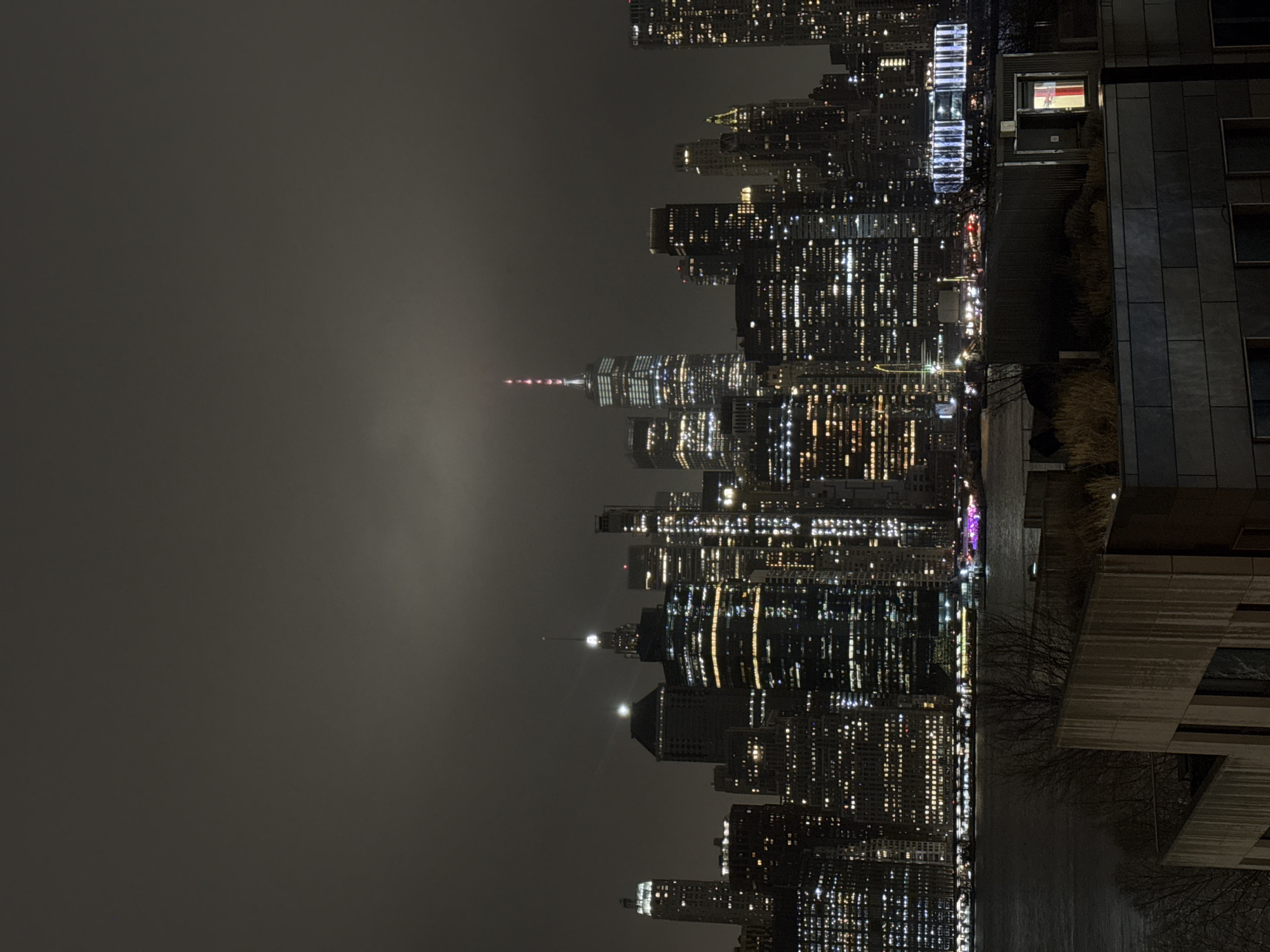 This image depicts a nighttime view of the New York City skyline on a hazy or foggy night. The buildings are brightly illuminated, with many visible lights from individual windows. The tallest building has a lit antenna on top. A river or body of water is located in front of the buildings. In the foreground, a corner of a building is visible, suggesting the photo was taken from a higher vantage point. The image is rotated, with the sky appearing on the left.