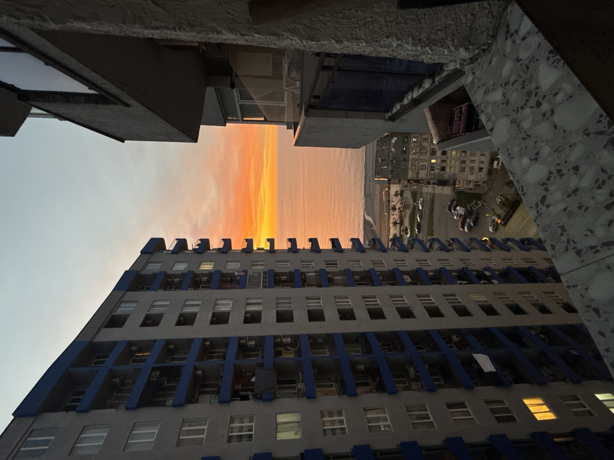 The image, taken from an elevated perspective, presents a view between two buildings in Batumi, Georgia, during what appears to be sunset. The lower building is a high-rise apartment structure with a gray facade and blue vertical architectural elements. Its balconies are visible, some with laundry hanging out to dry. The windows are mostly dark, though a few are lit. Between the two buildings, a panoramic view opens to the Black Sea, reflecting the orange hues of the setting sun. Across the sea, at a distance, sits another building. Several cars are parked along the road next to the building. The sky transitions from a light blue to a vibrant orange. The building from where the photo was taken has a balcony with a marble-like railing visible in the frame.
