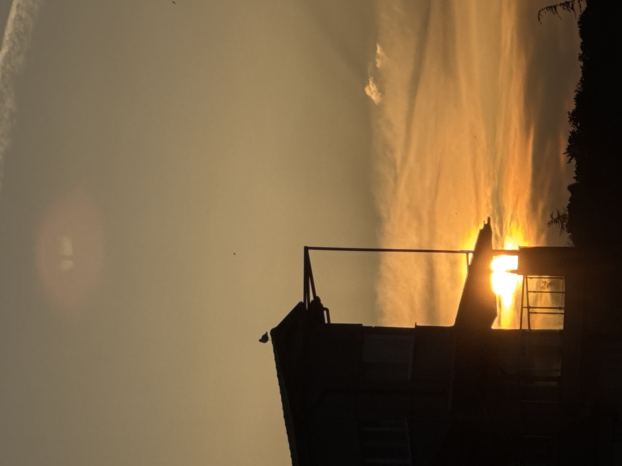 The image shows a silhouette of a building against a sunset. The sky is a mix of light orange and grey, with some clouds visible. The building has a pitched roof and a vertical structure. The sun is setting behind the building, creating a bright light. The location is unknown and is not identifiable as Tsarevo, Bulgaria. There are no people present in the image.