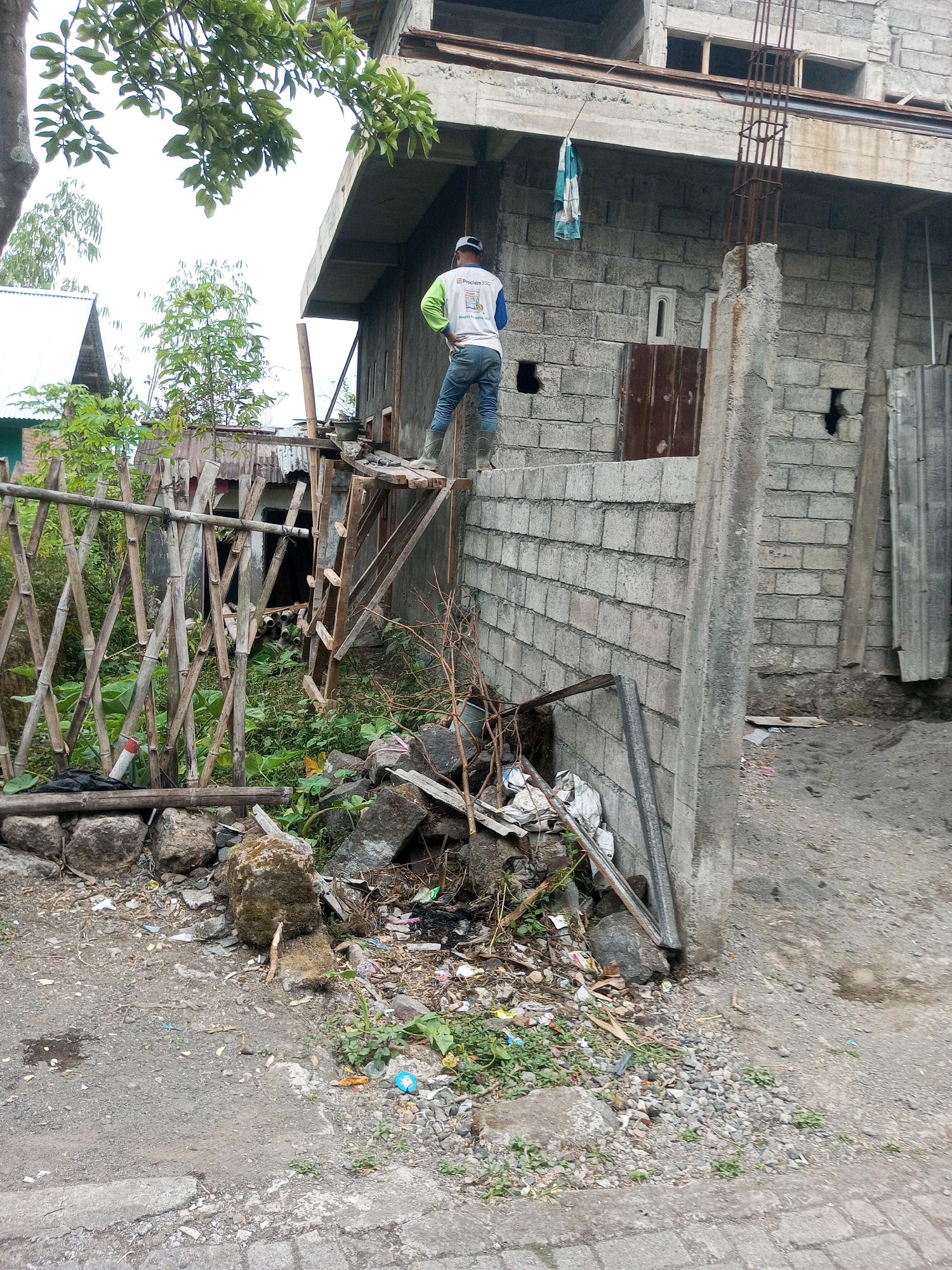The image depicts an outdoor scene in Andongsili, Indonesia, featuring a partially constructed building and its surroundings. A male individual wearing a white long-sleeved shirt with green accents, blue jeans, and dark boots stands on a temporary wooden platform attached to a cinder block wall. He is facing away from the camera, with his hands on his hips. The building, made of gray cinder blocks, appears to be under construction, with exposed rebar and concrete elements visible. To the left of the building, a fence constructed from bamboo poles creates a barrier. Scattered on the ground are debris, including rocks, trash, and pieces of wood. The ground surface is a mixture of dirt, gravel, and cracked concrete. Lush green vegetation, including trees and bushes, is present throughout the scene, contributing to the tropical environment. The sky is overcast, suggesting a cloudy day.