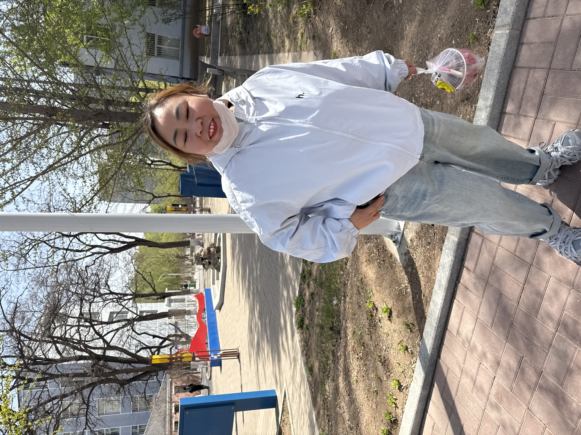 An Asian woman with brown hair, wearing a white jacket, blue jeans, and grey sneakers stands on a brick sidewalk. She is holding a plastic bag containing a cup with a straw. She is also wearing a white face mask. Behind her is a park-like area with trees, playground equipment (red, blue, and yellow), and buildings. The location is Zhongtun, China. A white metal rail is present to the left.