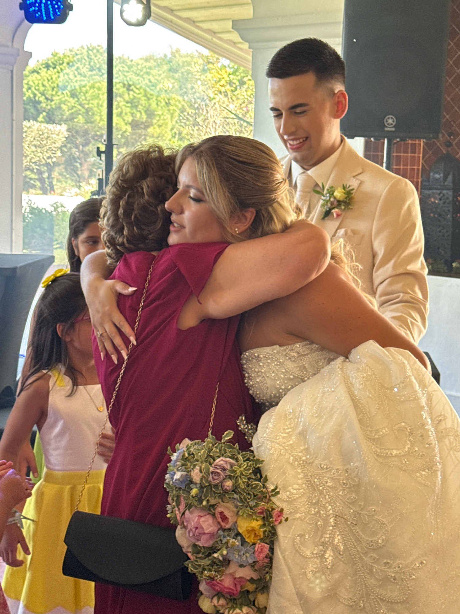 In the city of Mafra, Portugal, at what appears to be a wedding reception held indoors with natural light filtering in from an outdoor setting, a bride in a heavily beaded white wedding gown is embracing a woman wearing a burgundy dress. The bride's bouquet, a mix of pink, purple, and blue flowers, is visible near the woman's shoulder. The groom, in a cream-colored suit and boutonnière, stands behind them, smiling. In the background, two young girls are present; one in a yellow dress and the other partially obscured. The environment includes architectural elements like arches and what appears to be audio equipment, suggesting a celebration or event.