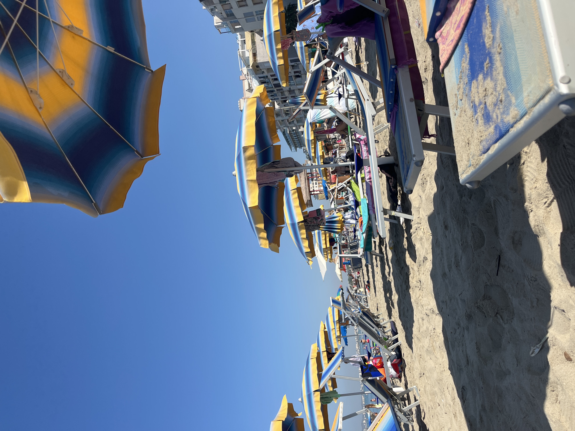 The image depicts a crowded beach with numerous lounge chairs and umbrellas set up on the sand. The umbrellas are predominantly blue and yellow striped, providing shade for the chairs. Several people are visible, some sitting on the chairs, while others are in the background, possibly swimming or walking along the shore. The ocean is visible in the distance, under a clear blue sky. Buildings line the background of the beach, suggesting an urban or resort area. The sand is light brown and appears well-trodden. The overall scene conveys a typical summer day at a popular beach destination.