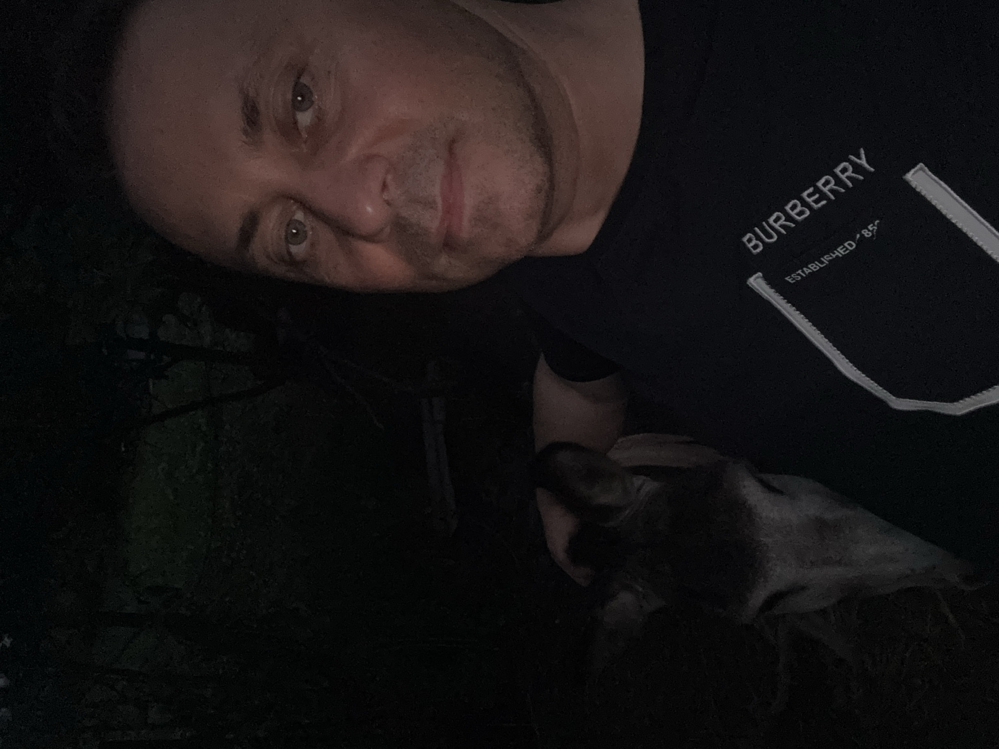 In the city of Tandzia, Georgia, a man is taking a selfie with a deer at night. The man is wearing a black t-shirt with a Burberry logo. The deer is looking at the camera, and the man is smiling. The background is dark, with trees and foliage visible. The image is taken in an outdoor setting.