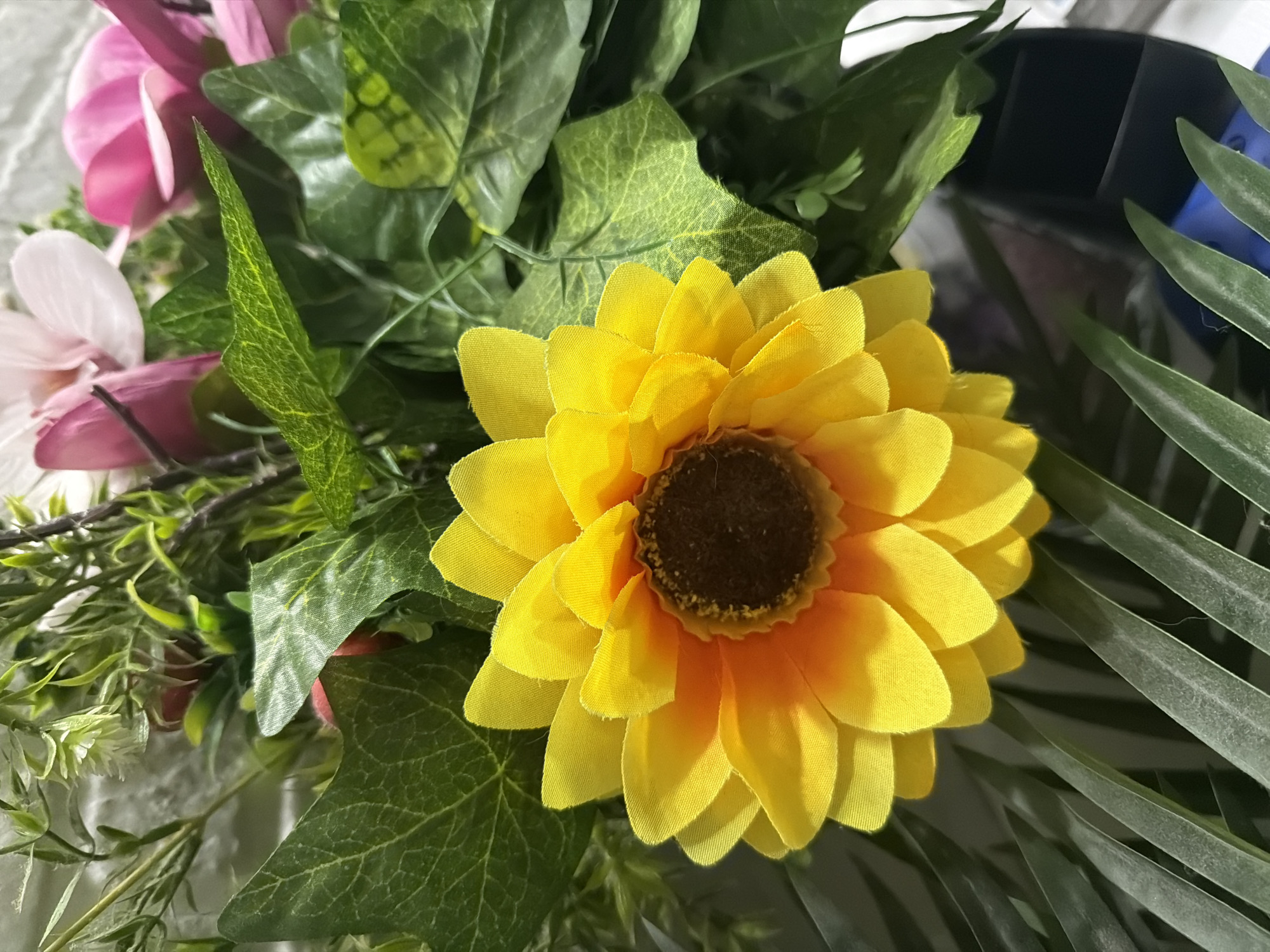 A close-up, eye-level, indoor shot focuses on a bright yellow artificial sunflower with a brown center. The sunflower is part of a floral arrangement that includes various green artificial leaves and some pink and white artificial flowers in the background. The arrangement is placed against a textured white wall. The lighting is bright, highlighting the textures and colors of the artificial flowers and foliage. The scene appears to be a still life arrangement, possibly for decorative purposes, and there is no indication of it being located in Tbilisi, Georgia, or any other specific city.