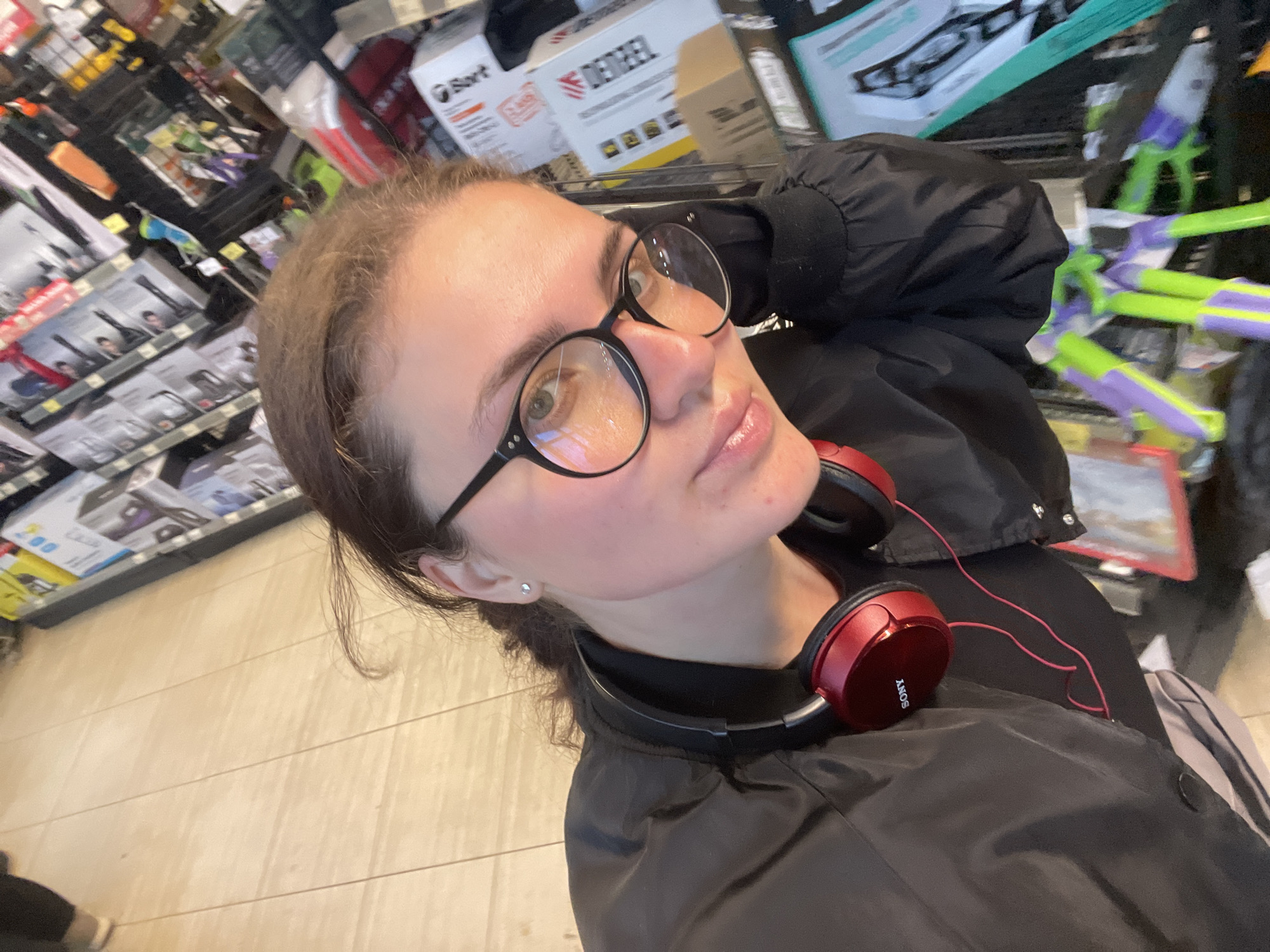 A young woman wearing round, black-rimmed glasses and red Sony headphones around her neck takes a selfie in a retail environment. She is dressed in a dark-colored jacket. Shelves filled with various products are visible in the background. The location is likely a retail store in Molokovo, Russia, based on other associated data. The floor has light-colored wood-look planks. There are water guns and boxes of DieReel on the shelf behind her.
