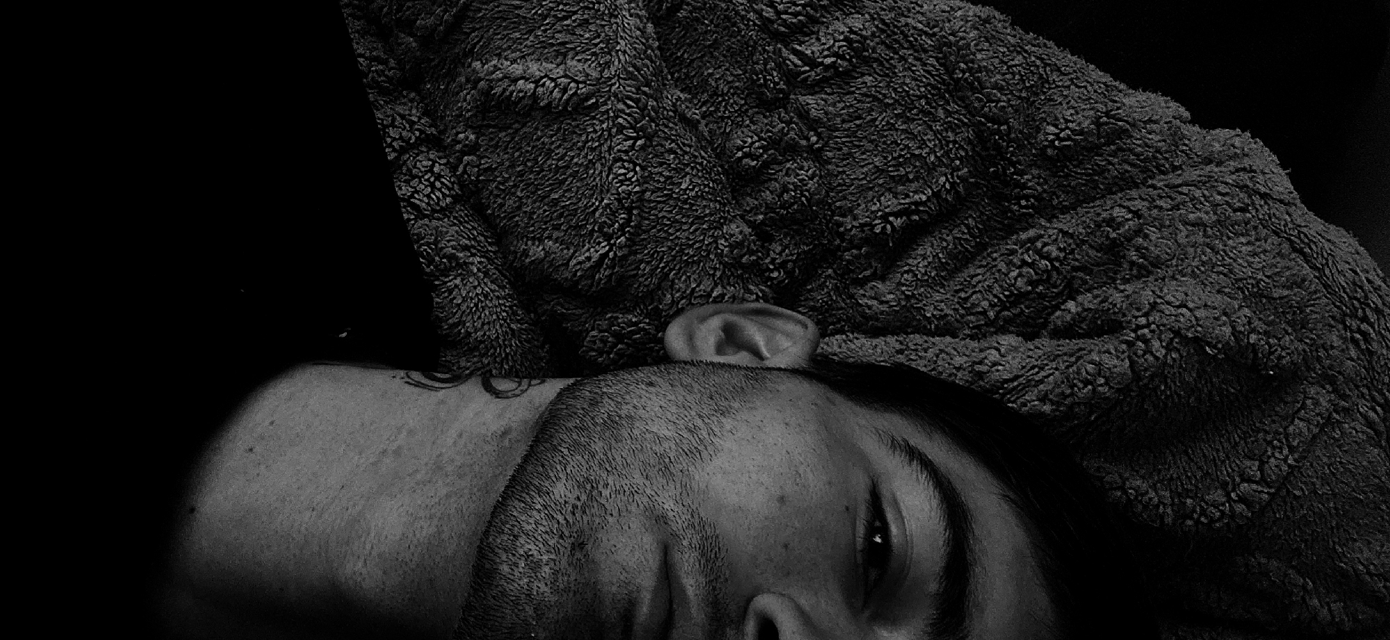 A black and white image depicts a man lying down, partially covered by a textured blanket. His head rests against the blanket, and his face is visible from the forehead to the upper lip. One eye is open, and a trimmed beard is visible. The blanket is dark and appears to be a fluffy or plush material. A portion of his neck is exposed, revealing what seems to be a tattoo. The background is dark and indistinct. There is no location information provided for the city of Plovdiv, Bulgaria.